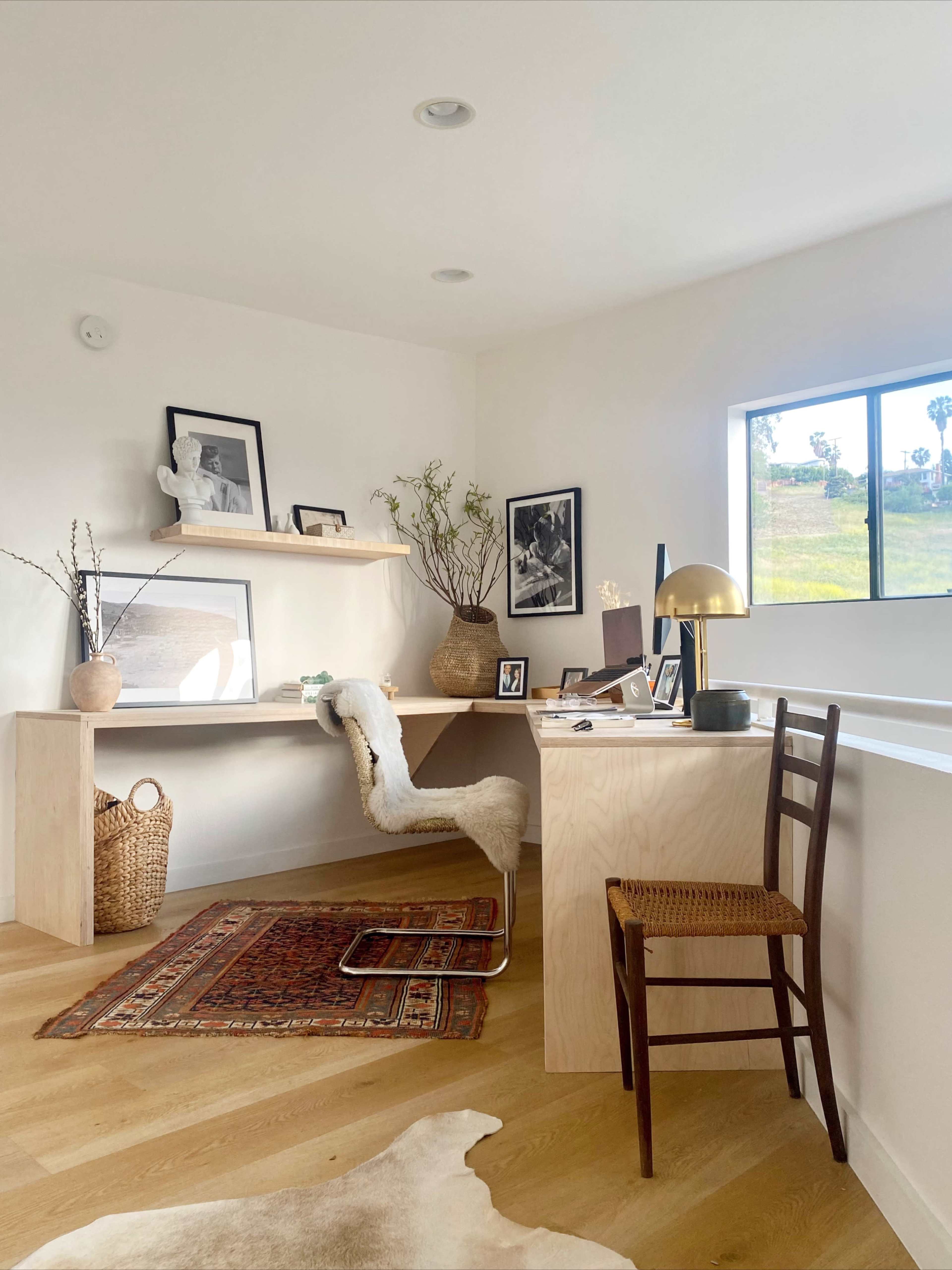 A stylish home office features a minimalist desk with a chair, decorative framed artwork on the walls, and a window overlooking a green landscape.