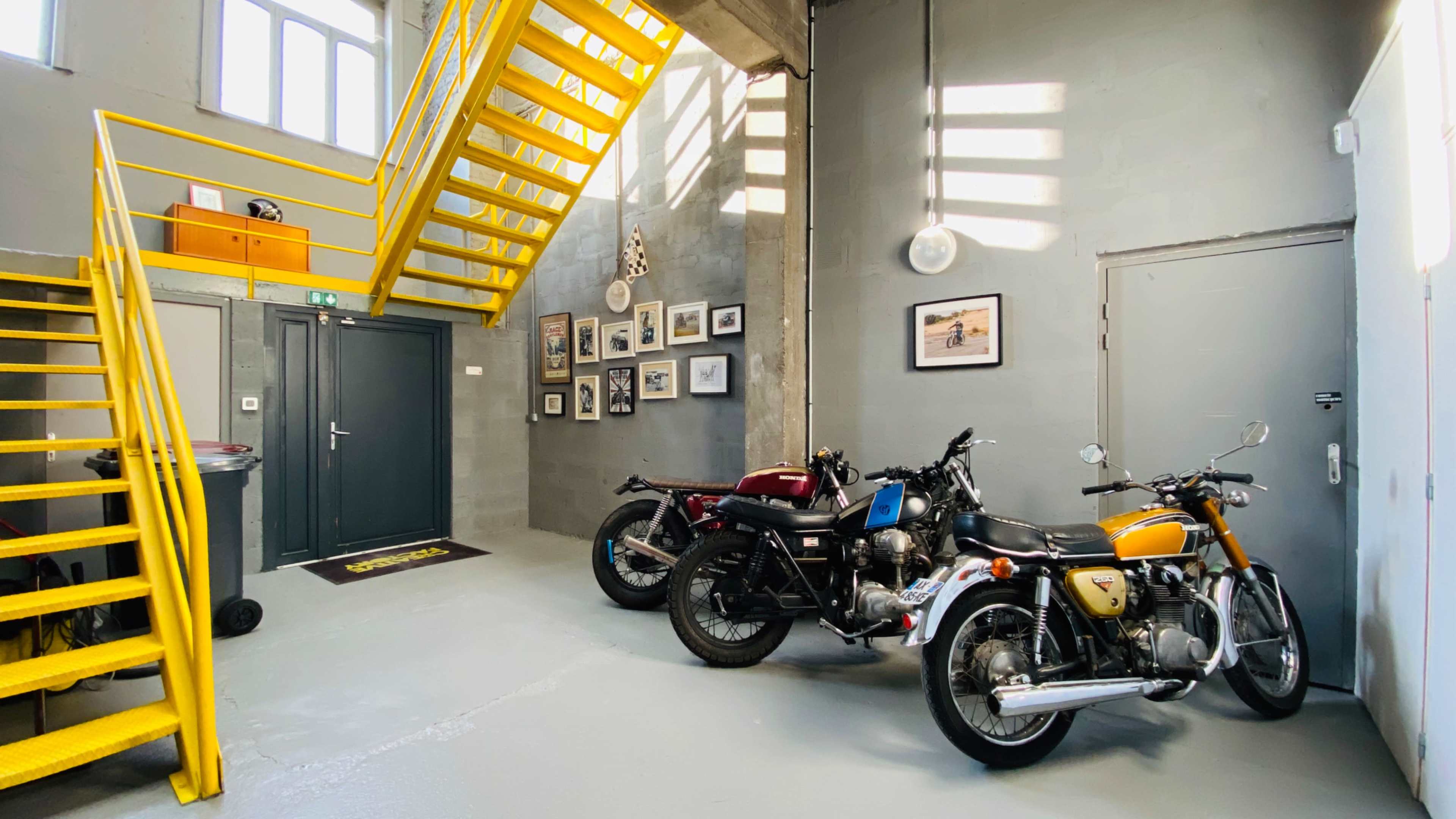 The image shows a modern garage space featuring three motorcycles parked near a gray wall, with a bright yellow staircase leading to an upper level.