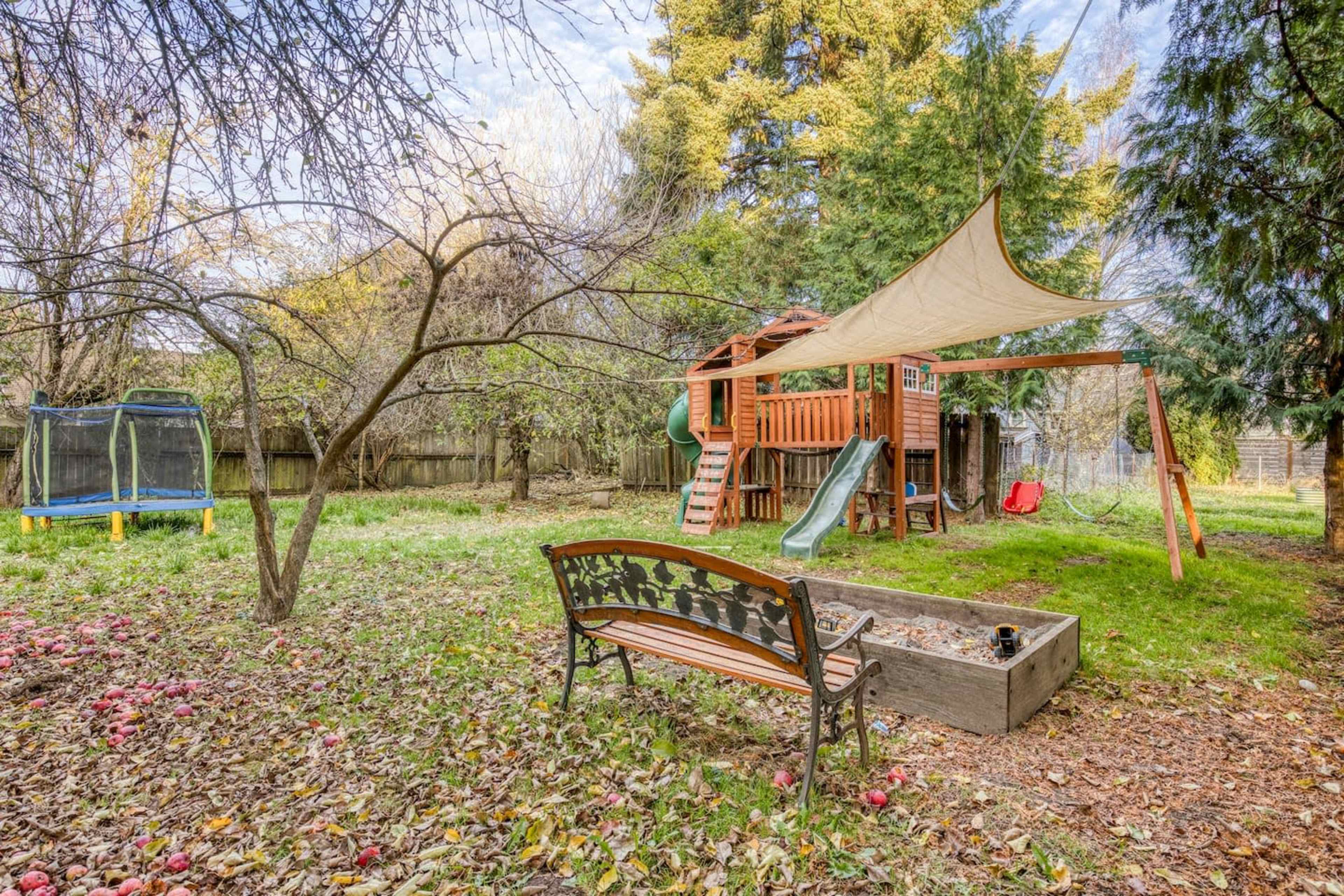 A backyard playground with a wooden play structure, a slide, swings, a sandbox, and a bench, surrounded by fallen leaves and apple remnants.