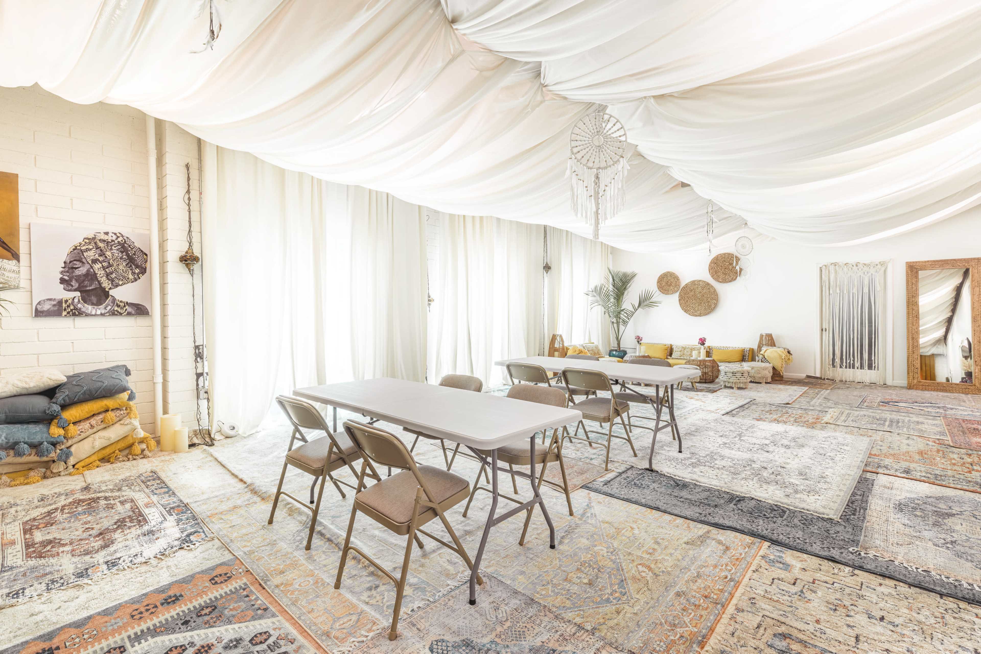 A spacious room with a white draped ceiling, several folding tables and chairs arranged, and various rugs on the floor.