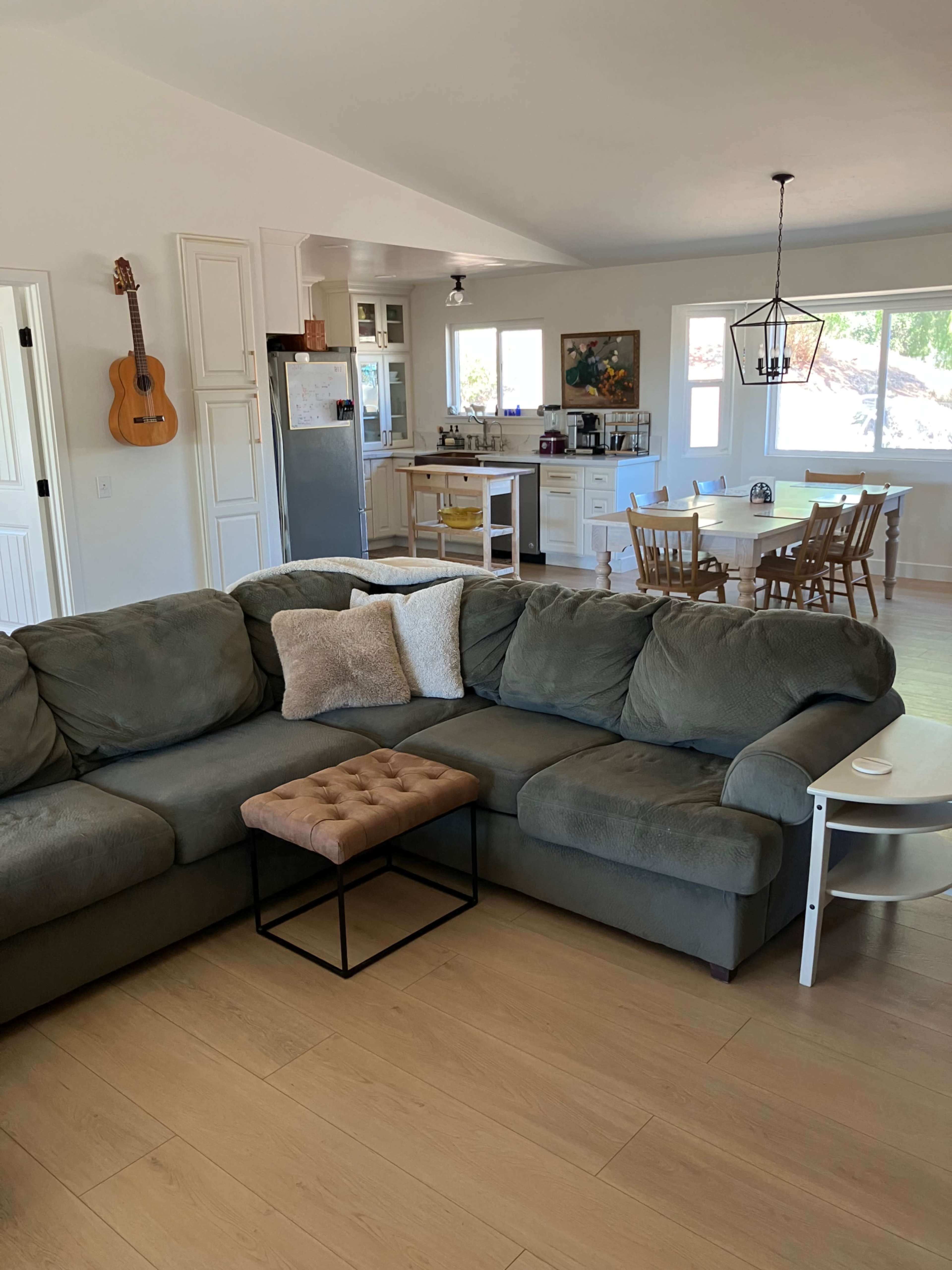 A cozy living room features a sectional sofa with a small ottoman in front, a dining area with wooden chairs and a table, and a kitchen visible in the background.