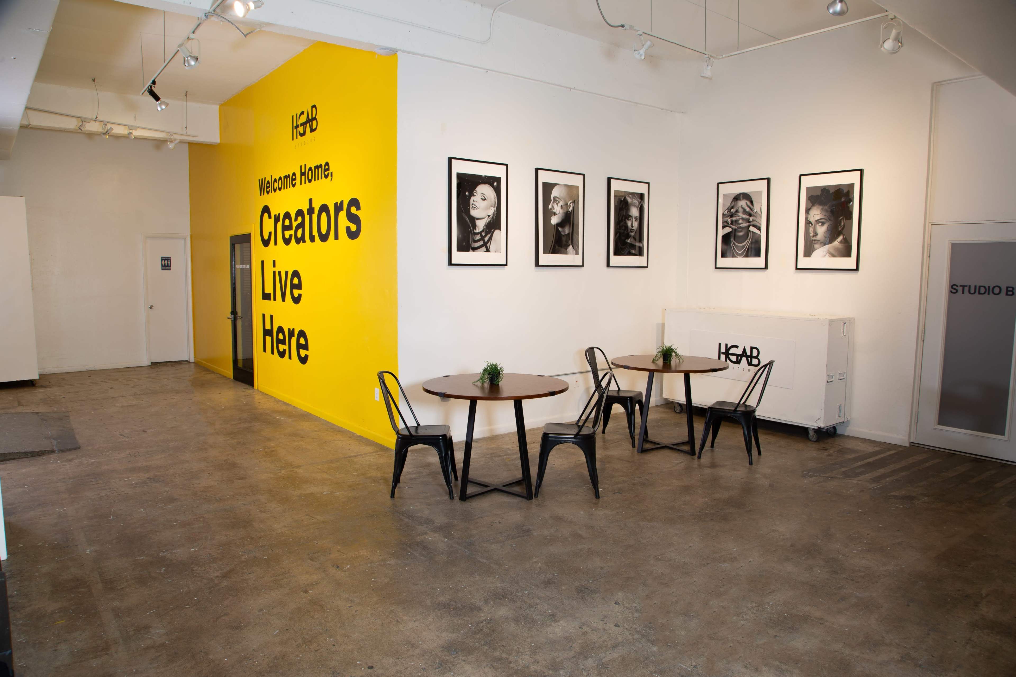 A bright yellow wall with black framed portraits, two round tables with chairs, and an entryway in a spacious interior.