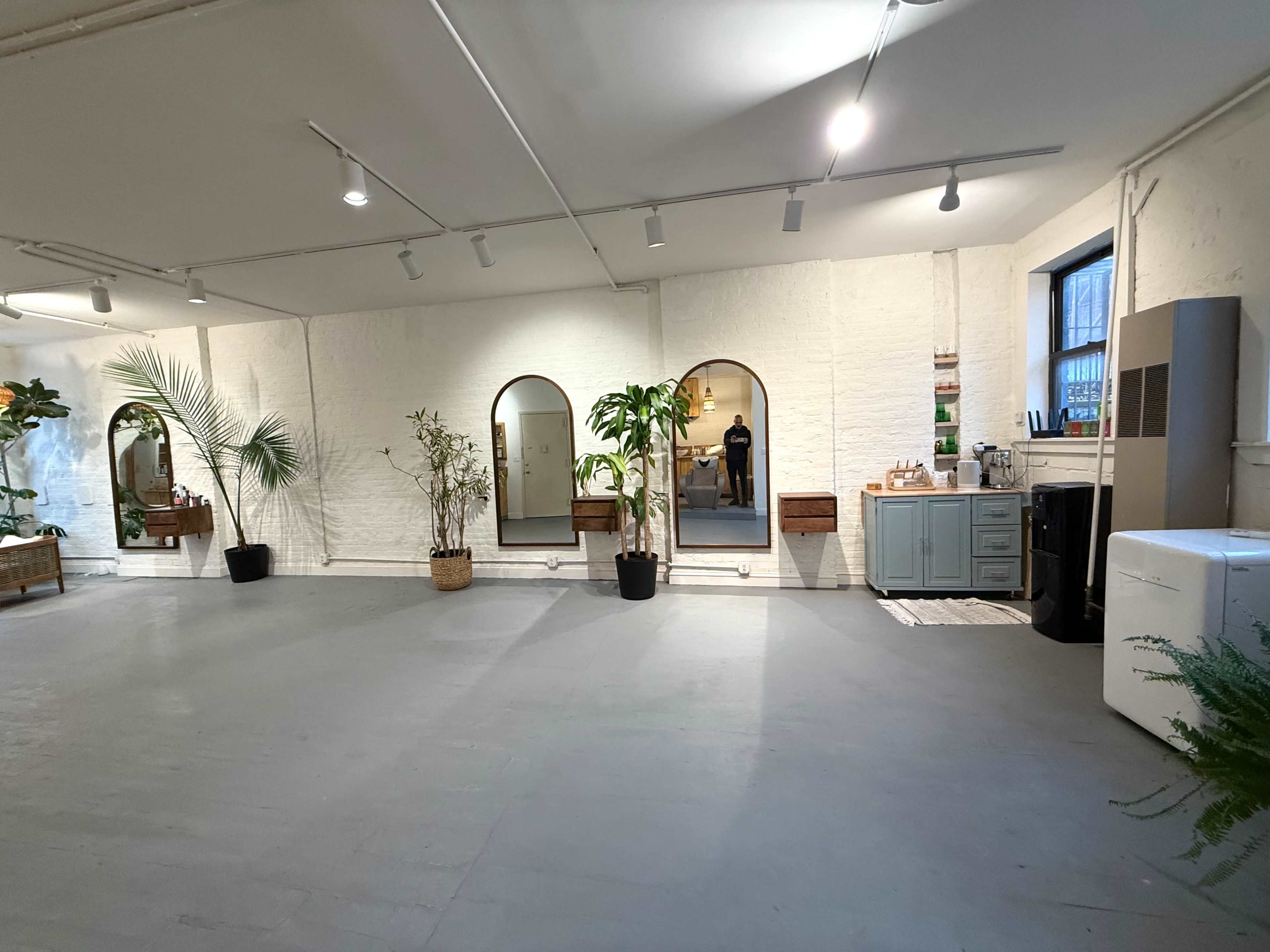 The image shows a spacious, well-lit room featuring plants, large mirrors, and a kitchenette area with various appliances against a textured white wall.