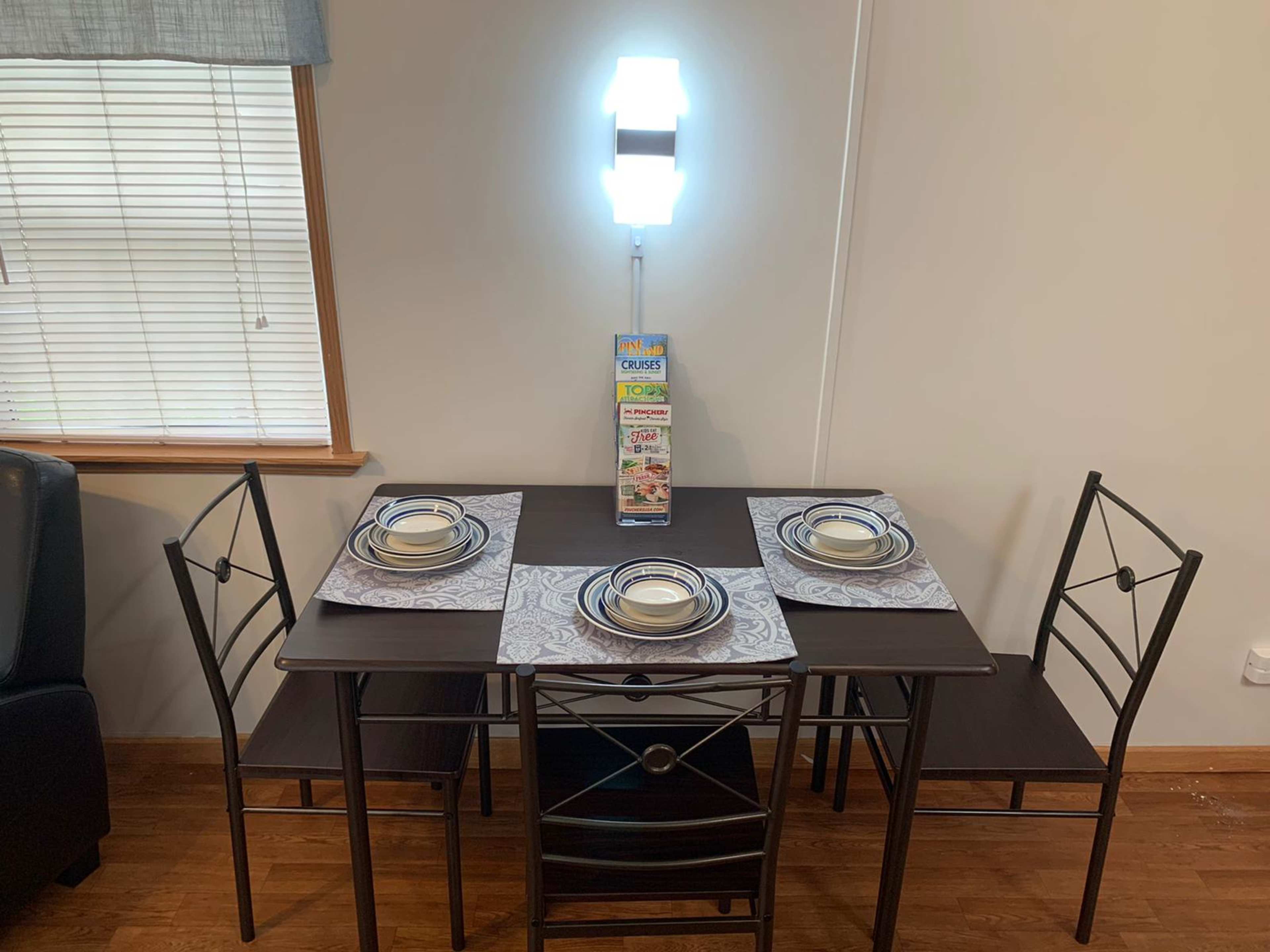 A small dining table set with four chairs, placemats, and plates, is positioned against a wall with a lamp and a display of snack boxes nearby.