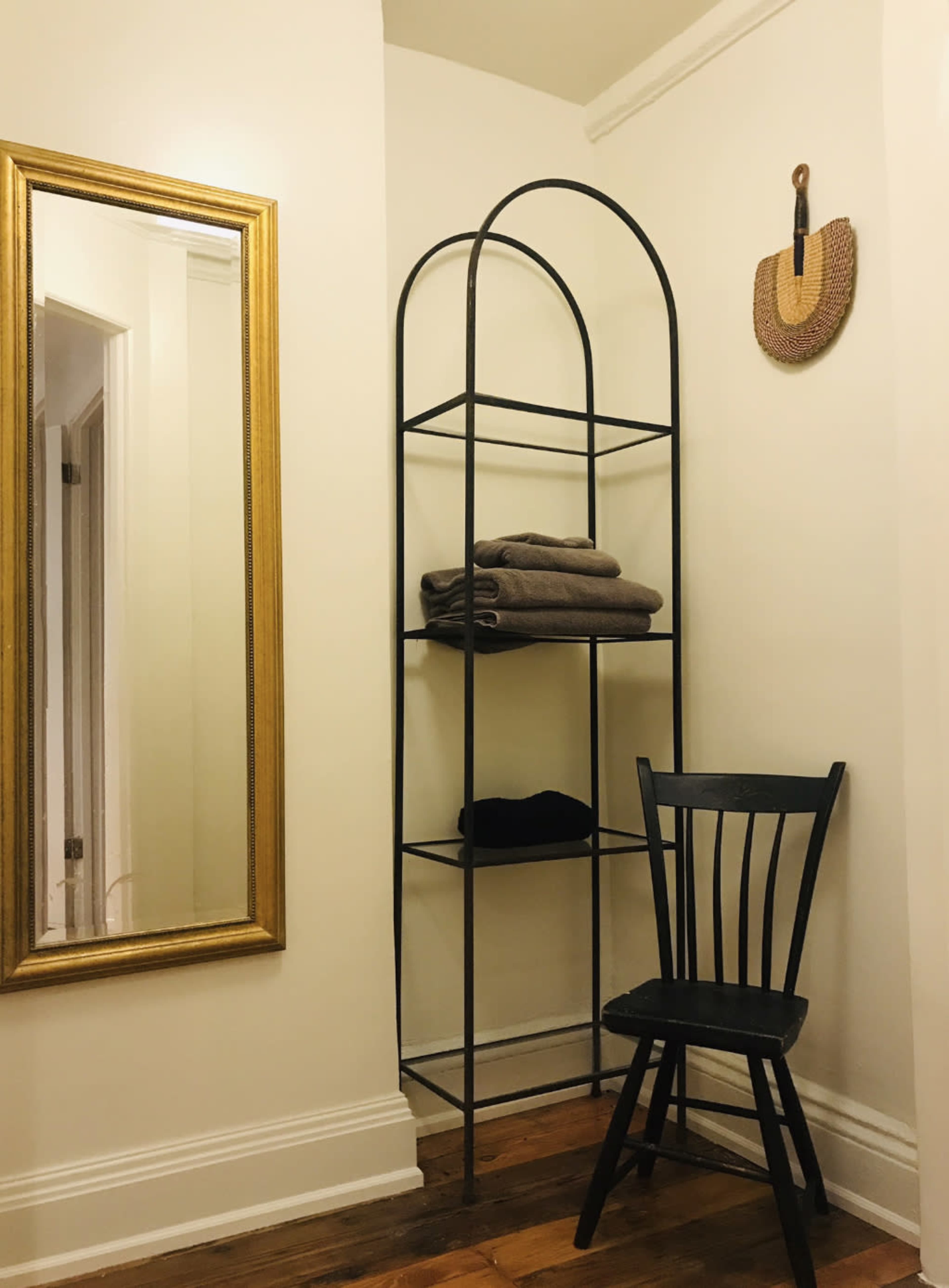 A black metal shelf with neatly stacked towels stands in a corner next to a mirror and a chair against a white wall.