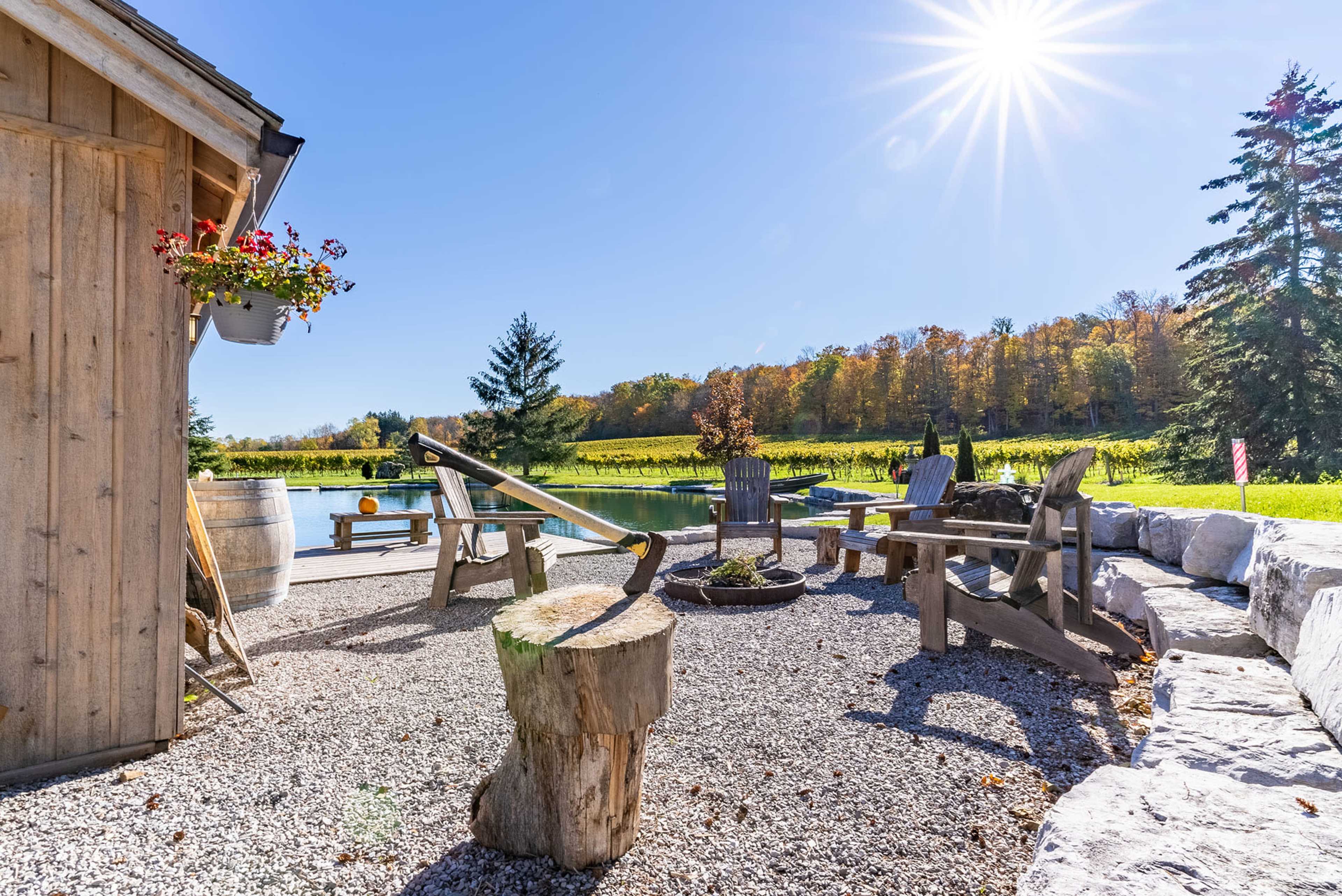 A gravel patio features wooden chairs and a fire pit, surrounded by trees and a vibrant landscape under bright sunlight.