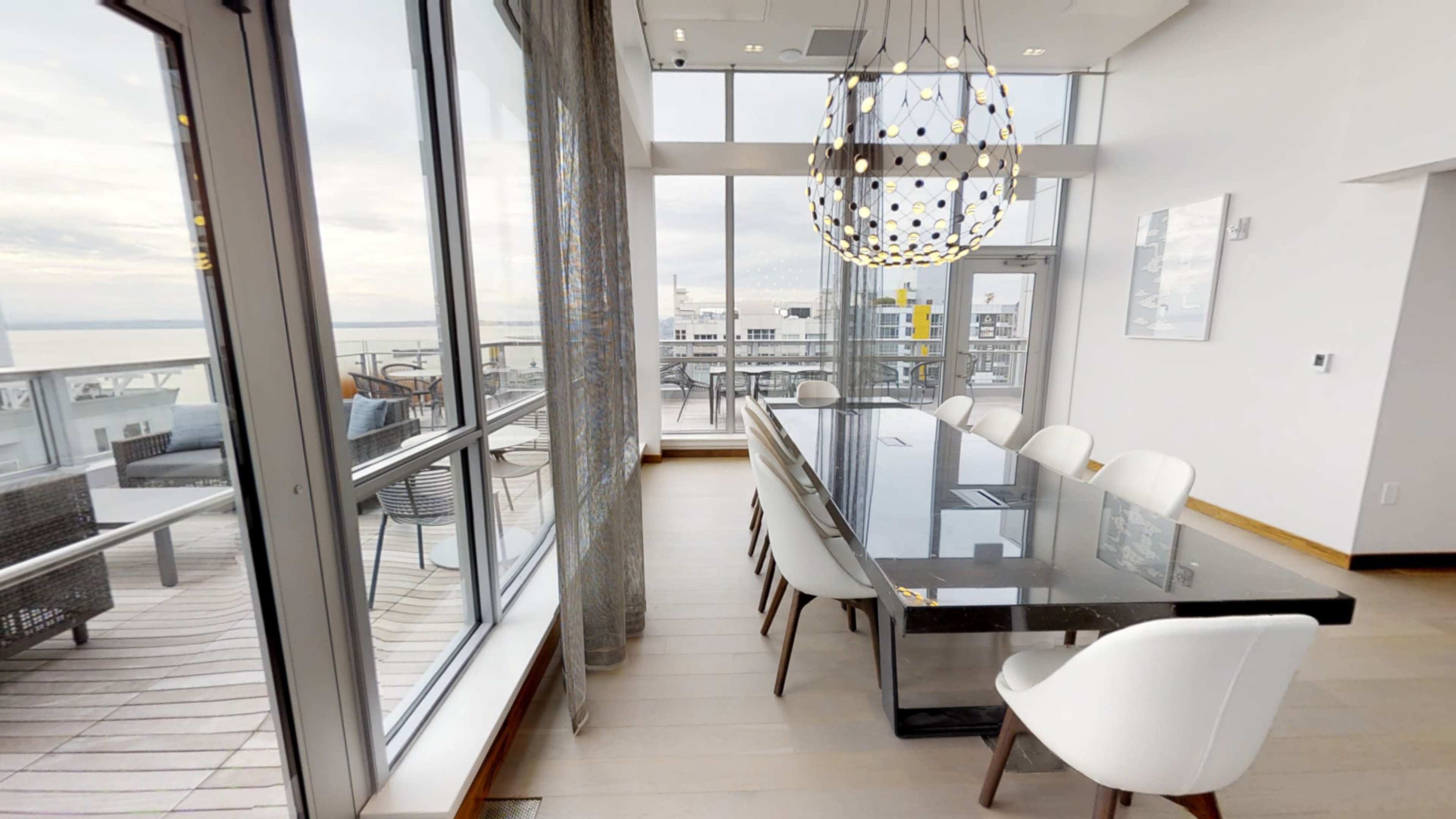 A modern dining room features a long glass table surrounded by white chairs, with large windows offering a view of a balcony and outdoor seating.