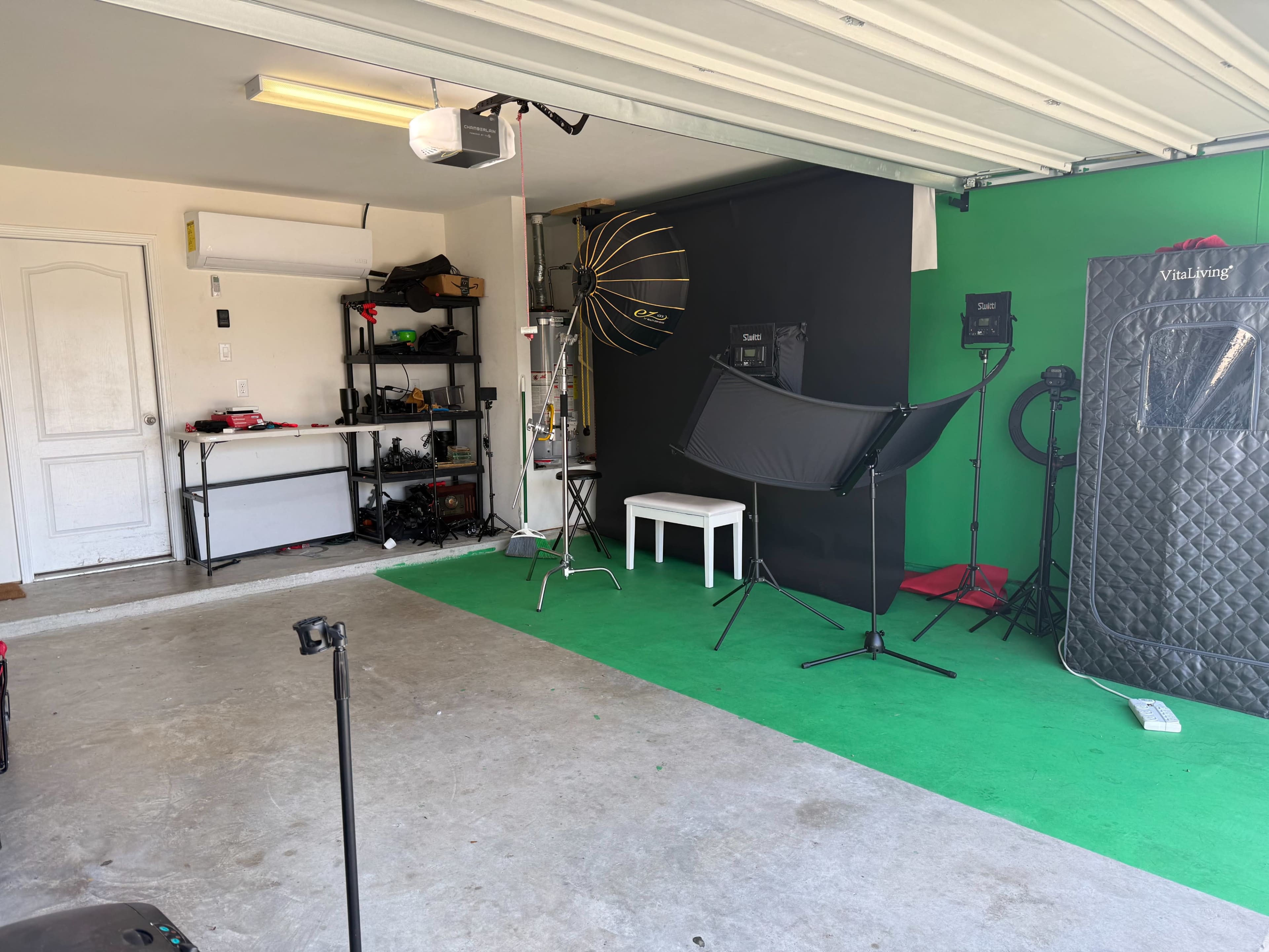 The image shows a garage studio setup featuring a green screen, lighting equipment, and a small table.