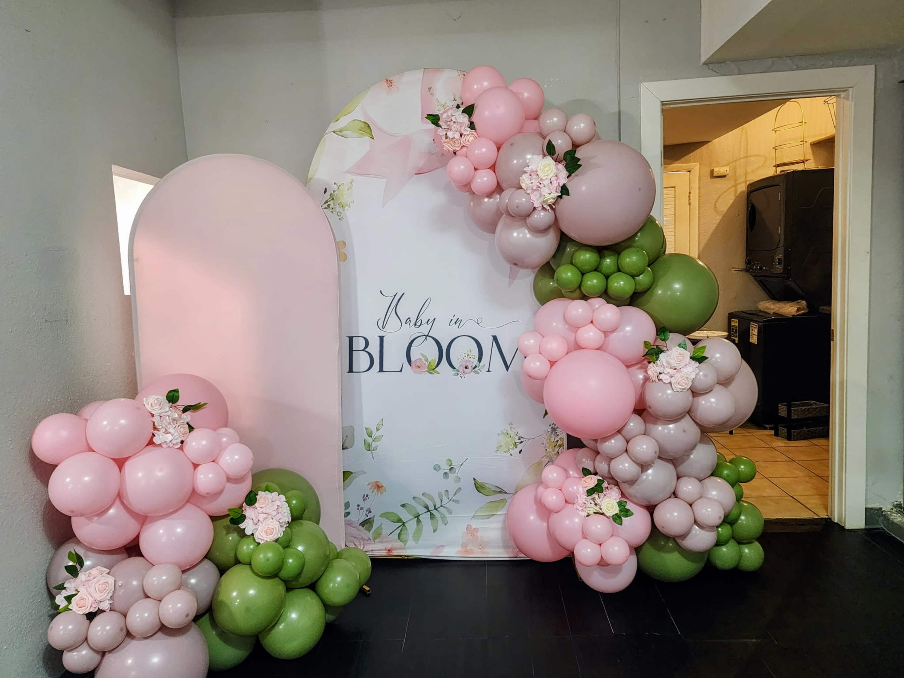 The image shows a decorative backdrop featuring a floral design with the text "Baby in Bloom," surrounded by arrangements of pink, green, and beige balloons.