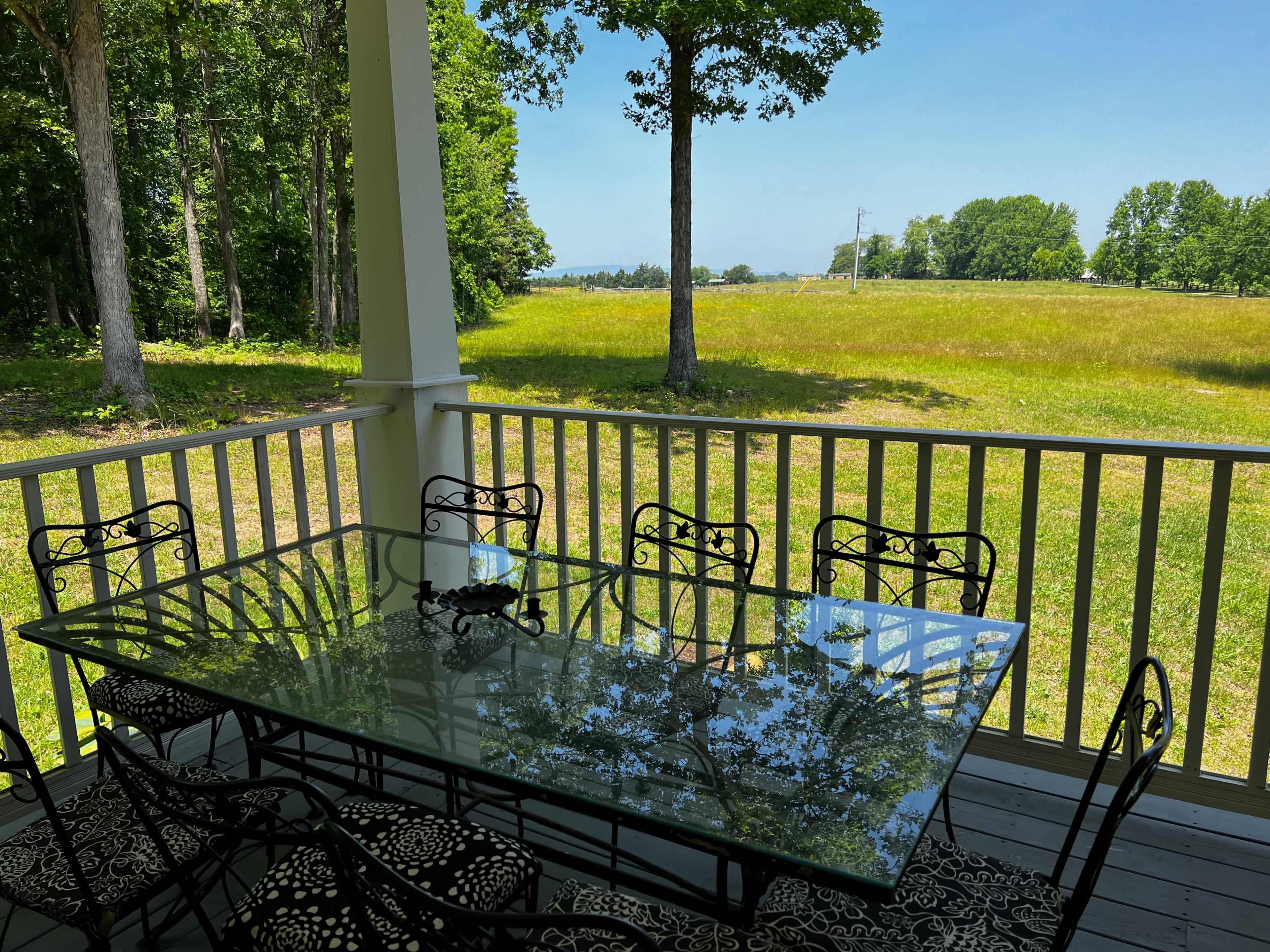 A glass dining table with metal chairs is positioned on a porch overlooking a green field and trees under a clear blue sky.