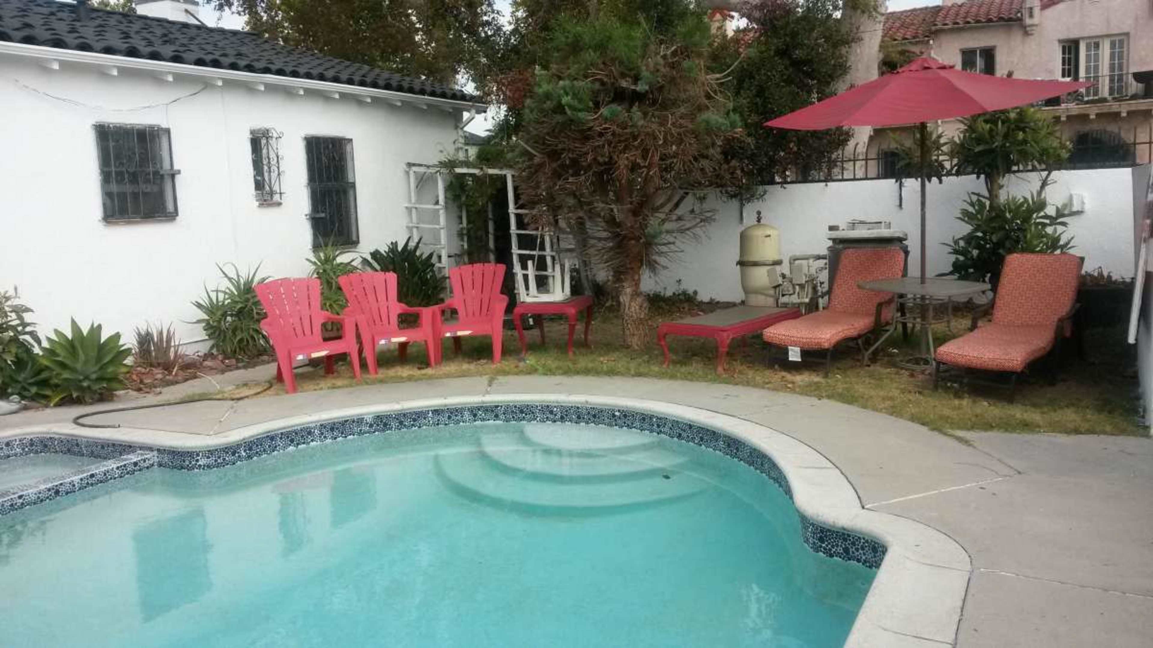 The image shows a swimming pool surrounded by red outdoor chairs, lounge chairs, and an umbrella in a backyard garden setting.