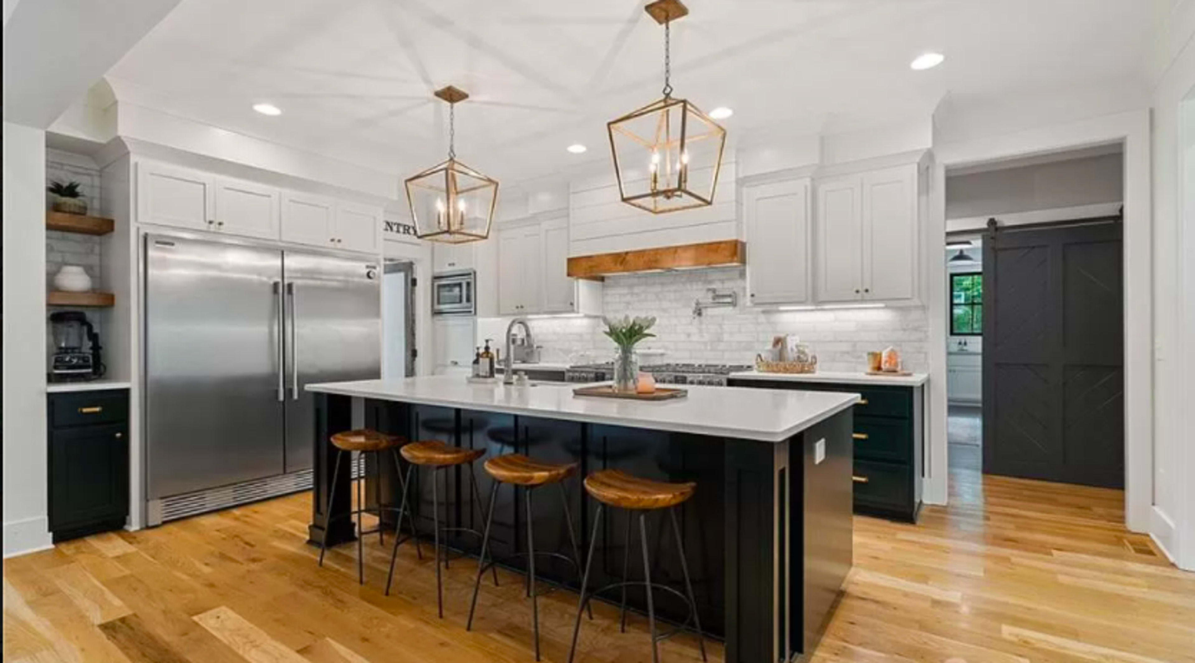 A modern kitchen features a central island with bar stools, stainless steel appliances, white cabinetry, and wooden accents on the countertops and ceiling lights.