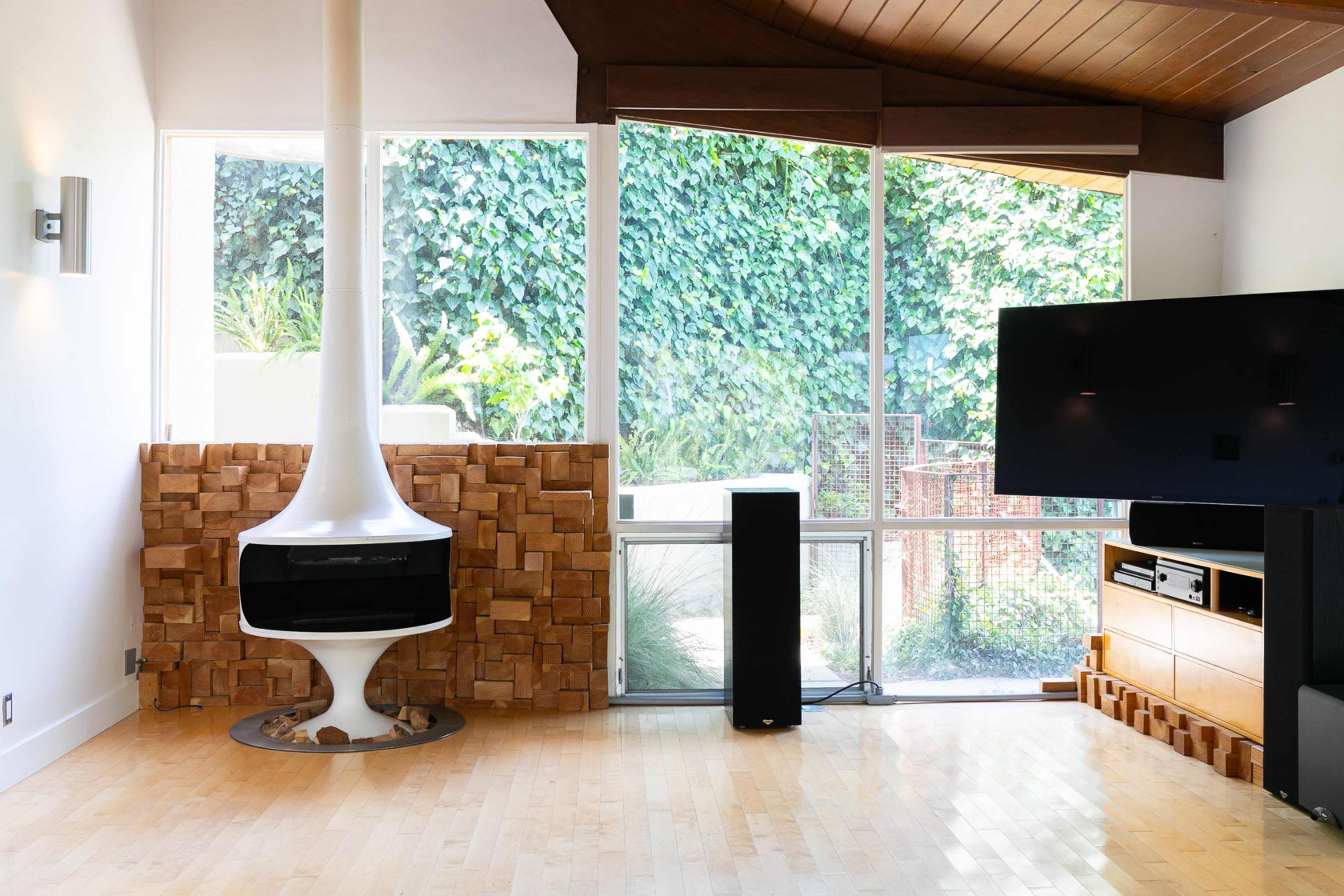 The image shows a modern living room featuring a white, curved fireplace, a large flat-screen TV, and wooden furnishings, all set against a backdrop of greenery visible through large windows.