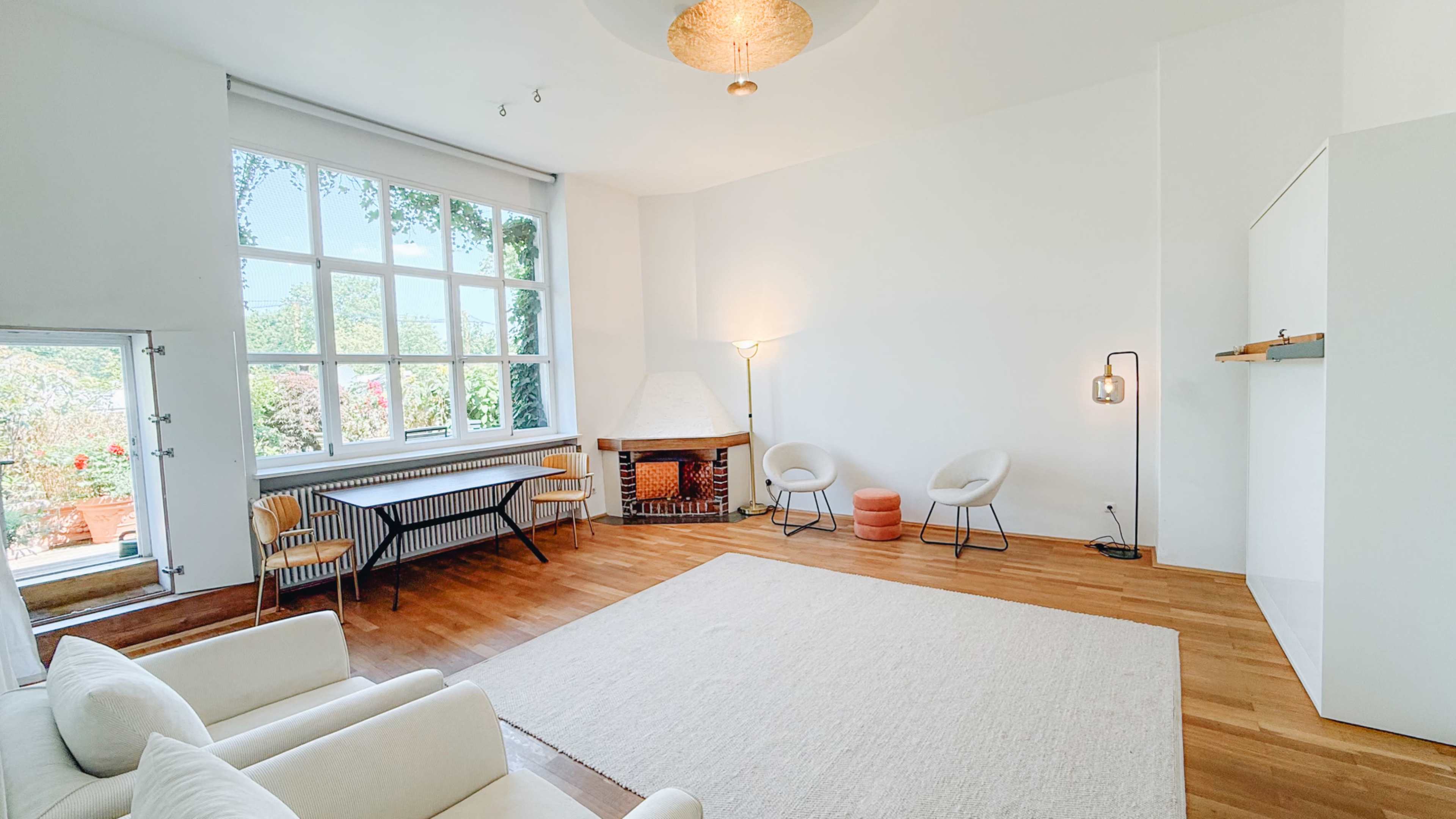 The image shows a bright, minimalistic living room with large windows, a fireplace, and two light-colored chairs facing an empty area on a wooden floor.