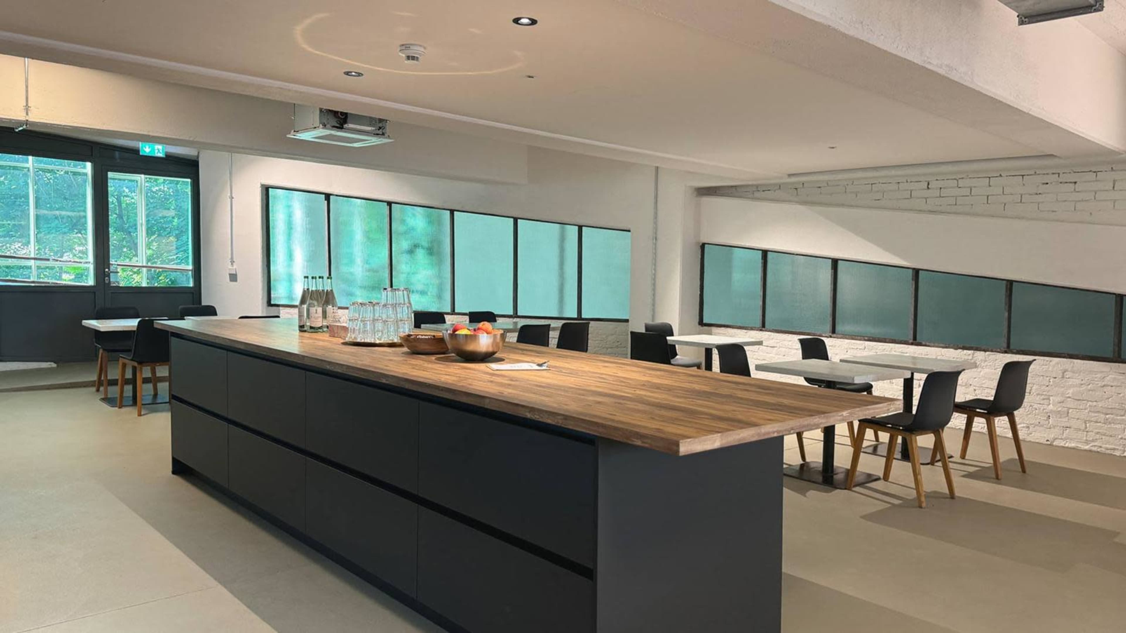 A modern kitchen with a large wooden island, black cabinetry, and several dining tables arranged in a well-lit space with large windows.