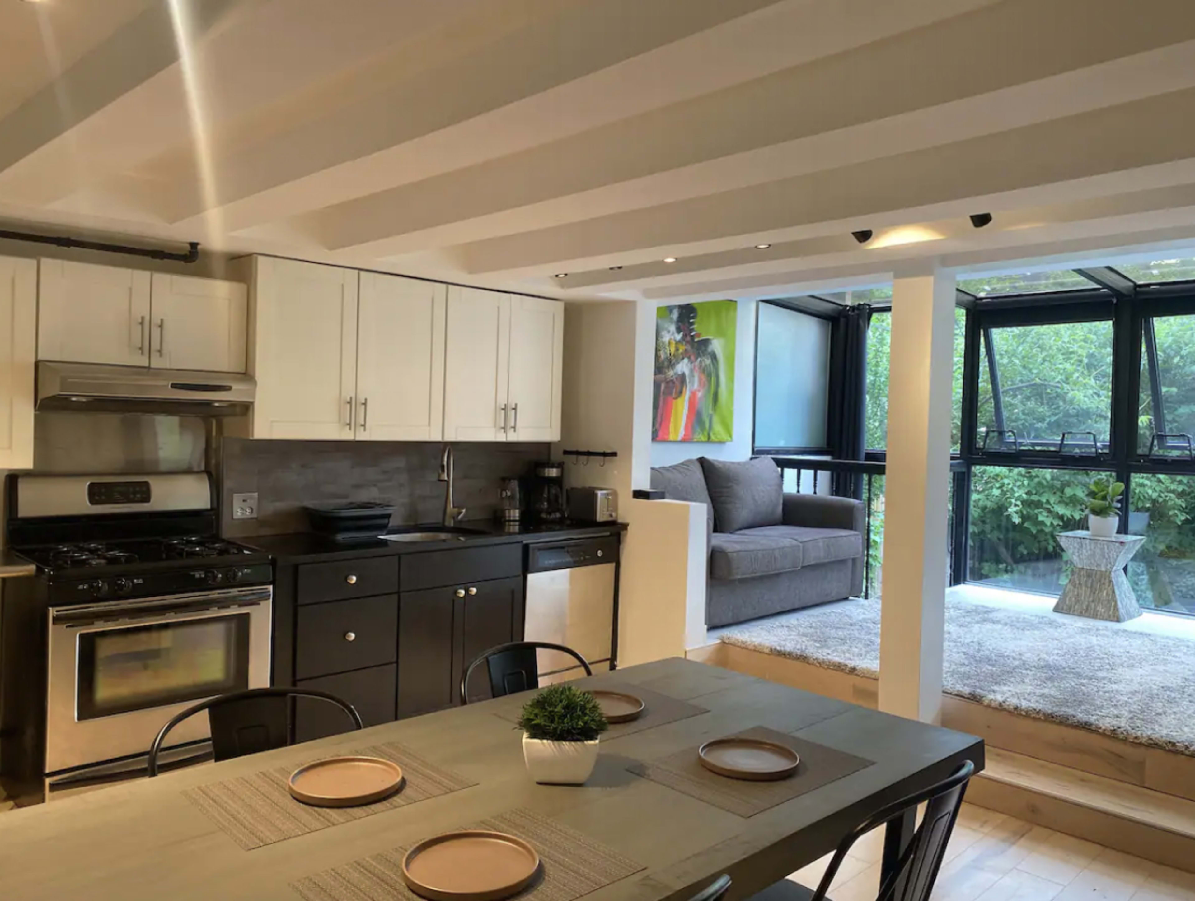 The image shows a modern kitchen with white cabinets and a black stove, adjoining a sunlit seating area with a large window and a soft rug.