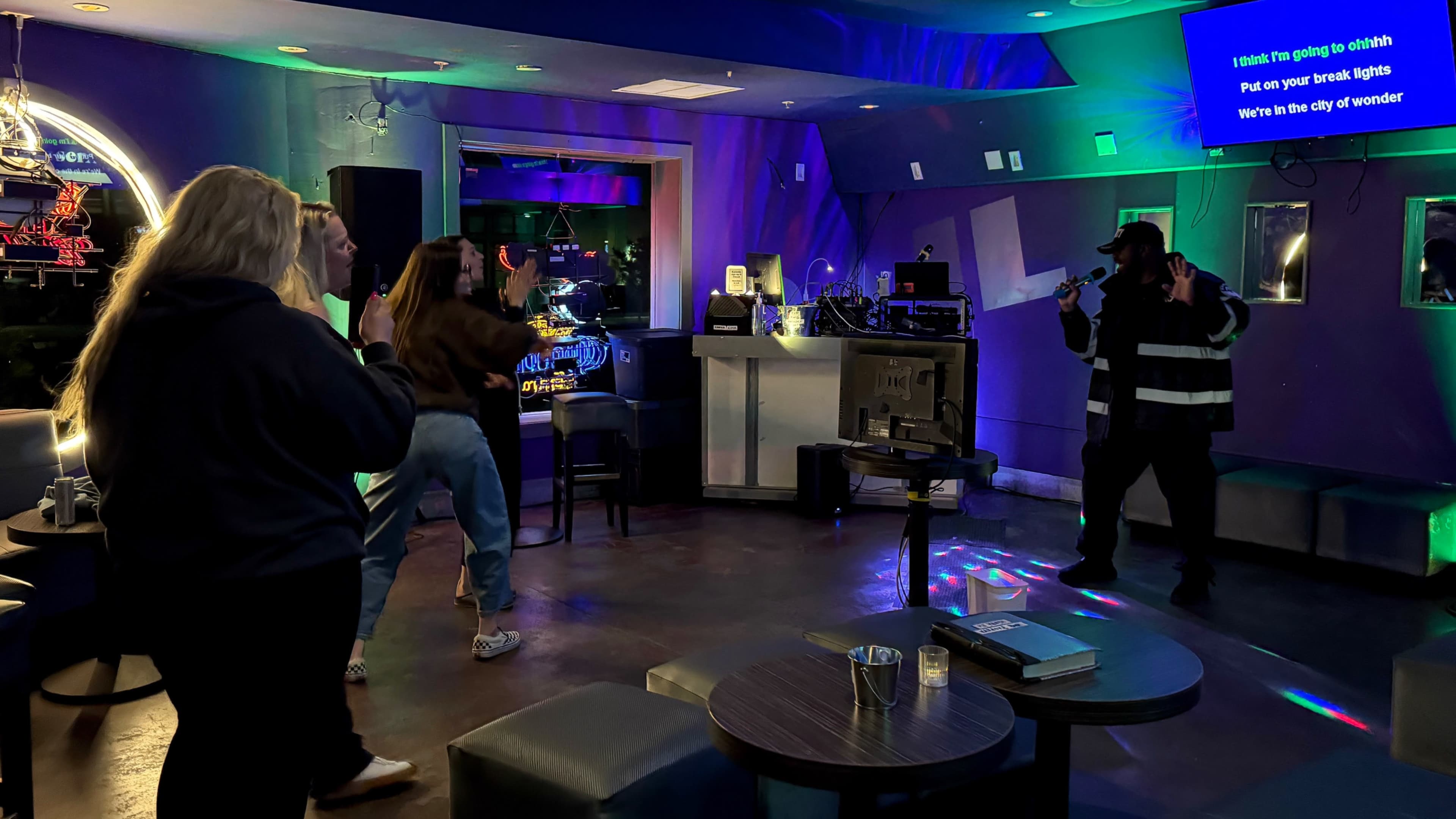 A group of people dances in a bar while a performer sings on stage with colorful lights illuminating the scene.