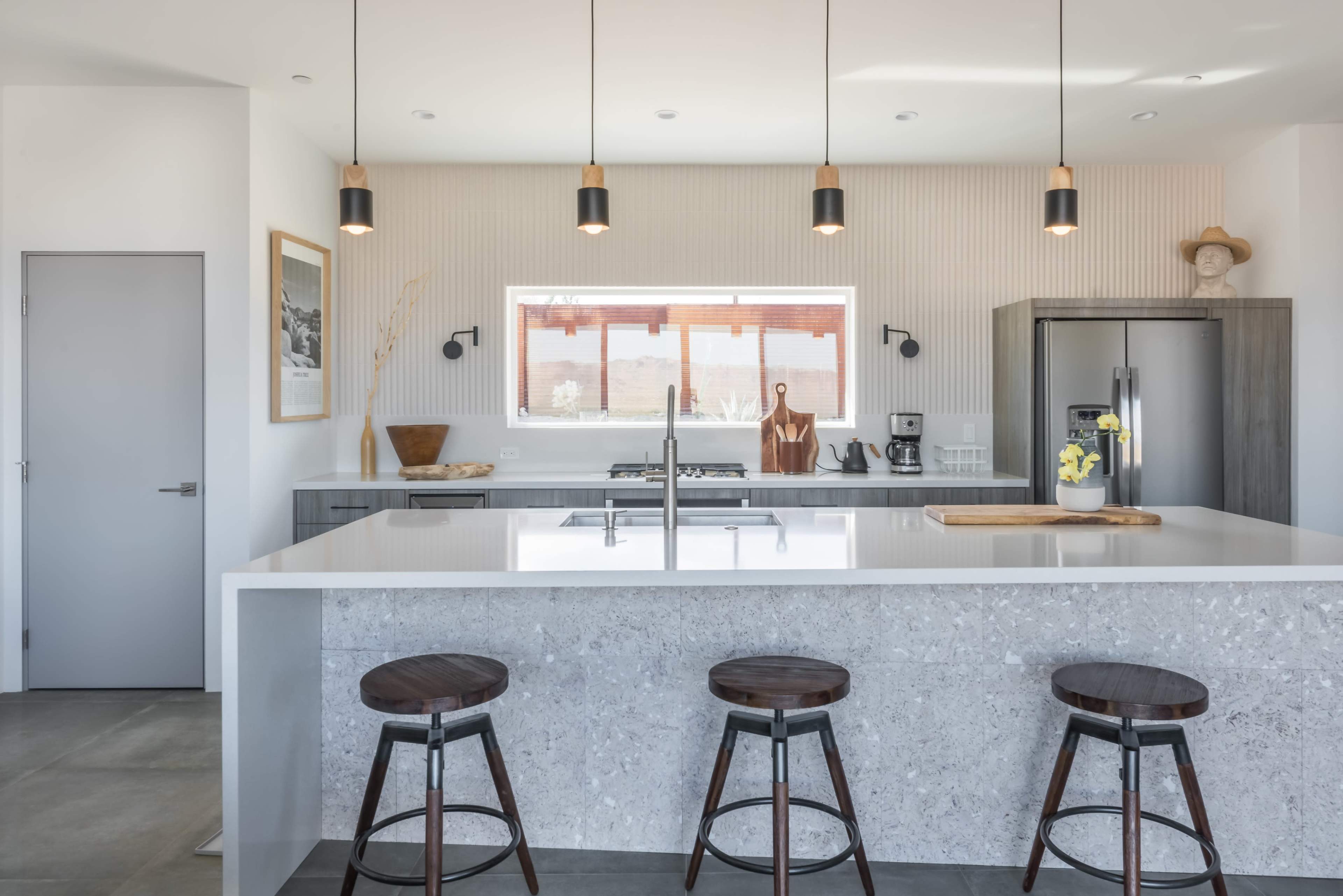The image shows a modern kitchen featuring a large island with three wooden stools, stainless steel appliances, and minimalist decor.