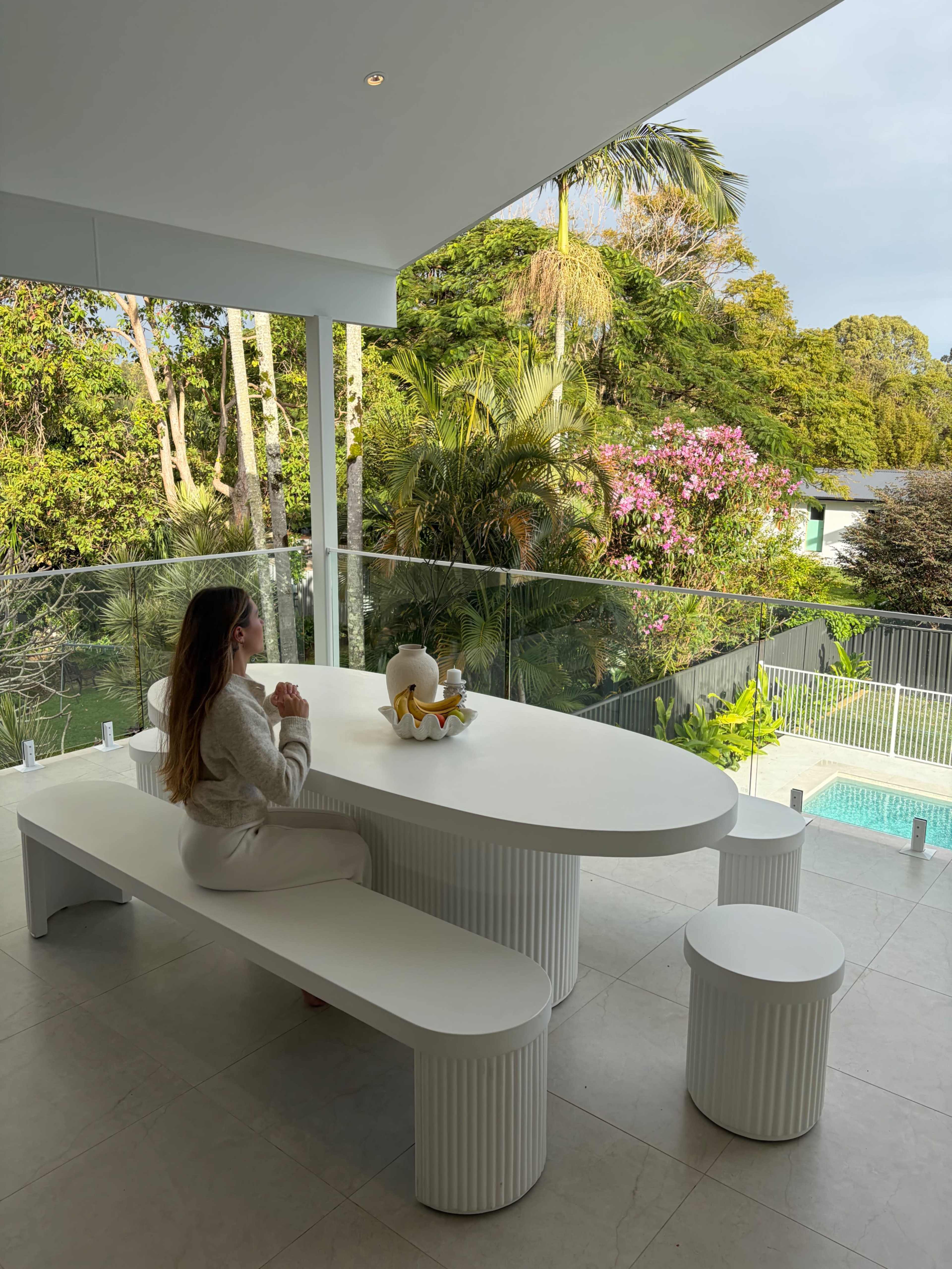 A woman sits at a modern white table on a covered patio overlooking a pool and lush greenery.