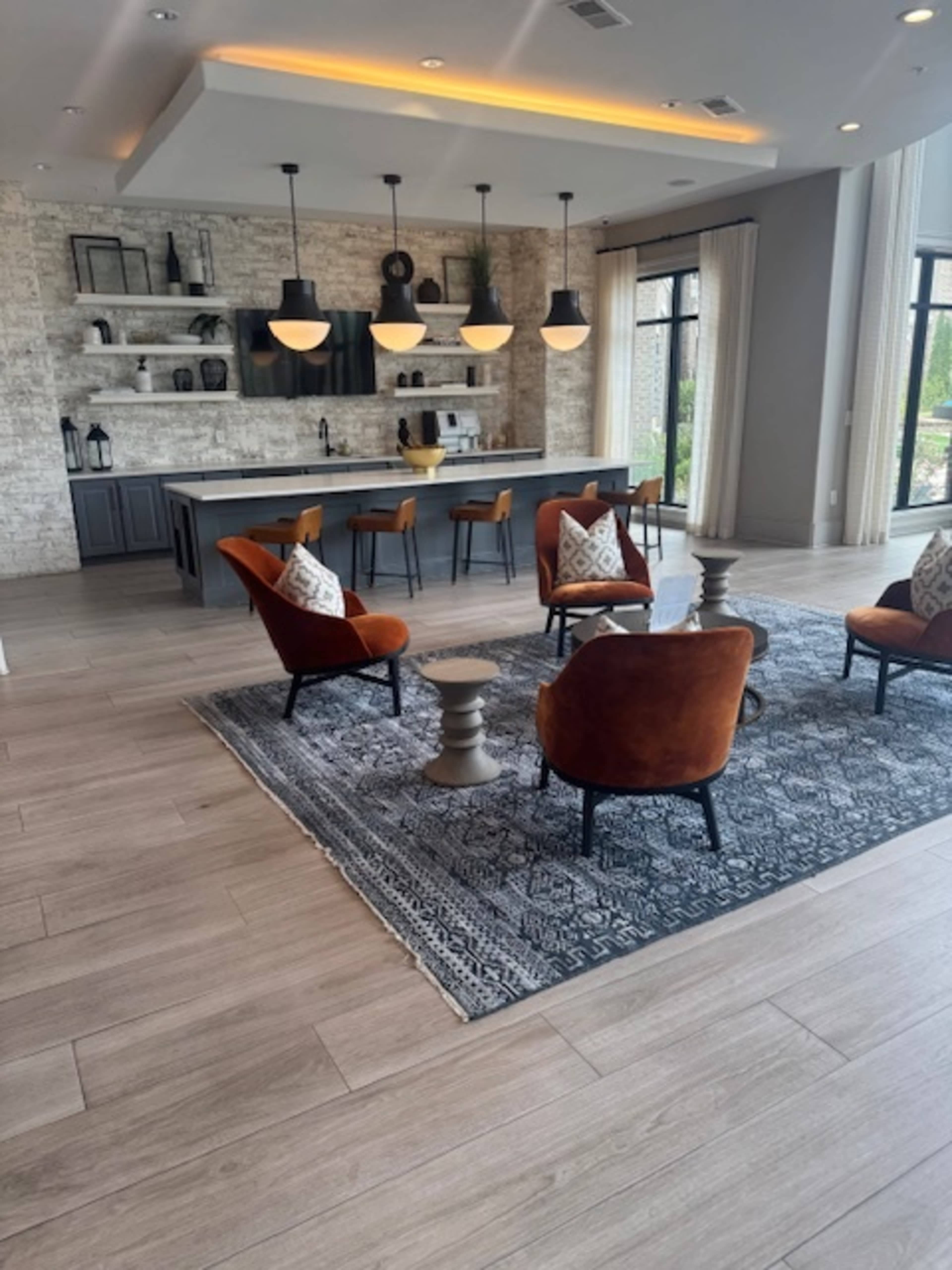 The image shows a modern living space featuring four orange chairs arranged around a circular table on a patterned rug, with a kitchen area and shelves in the background.