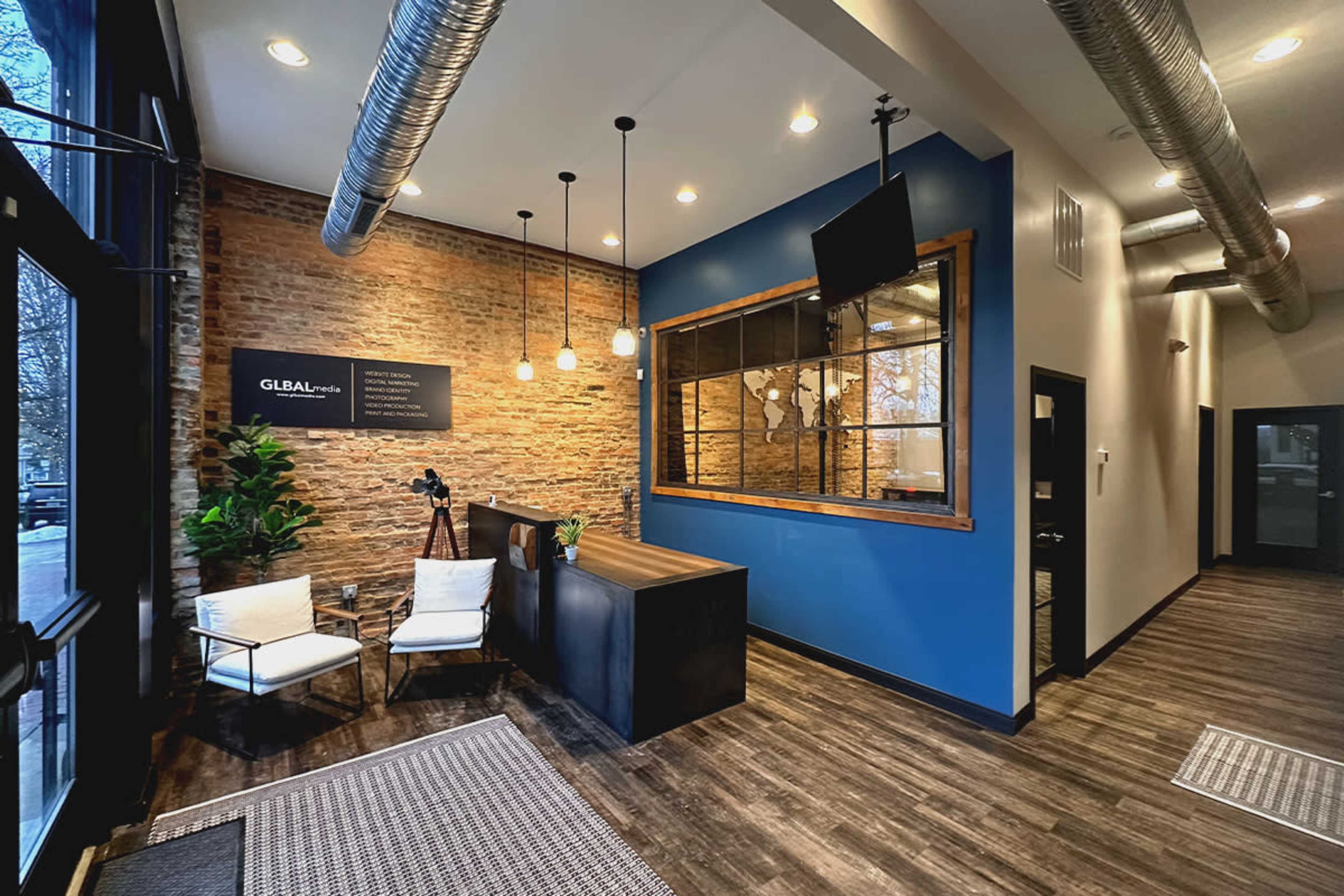 A modern reception area featuring a brick wall, a dark wooden reception desk, and two white chairs next to a tall plant.