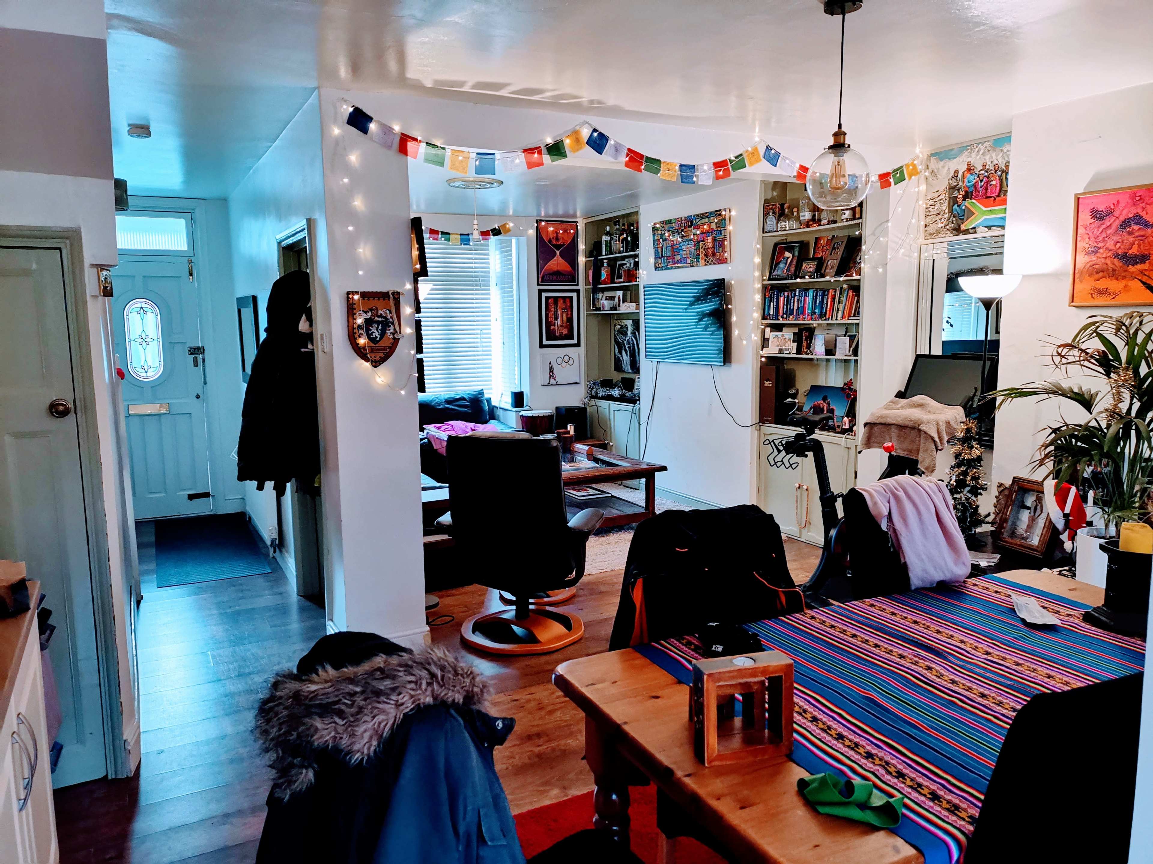 A bright, open living space with a dining table, a colorful banner hanging from the ceiling, and various furnishings, including a black office chair and shelves of books.
