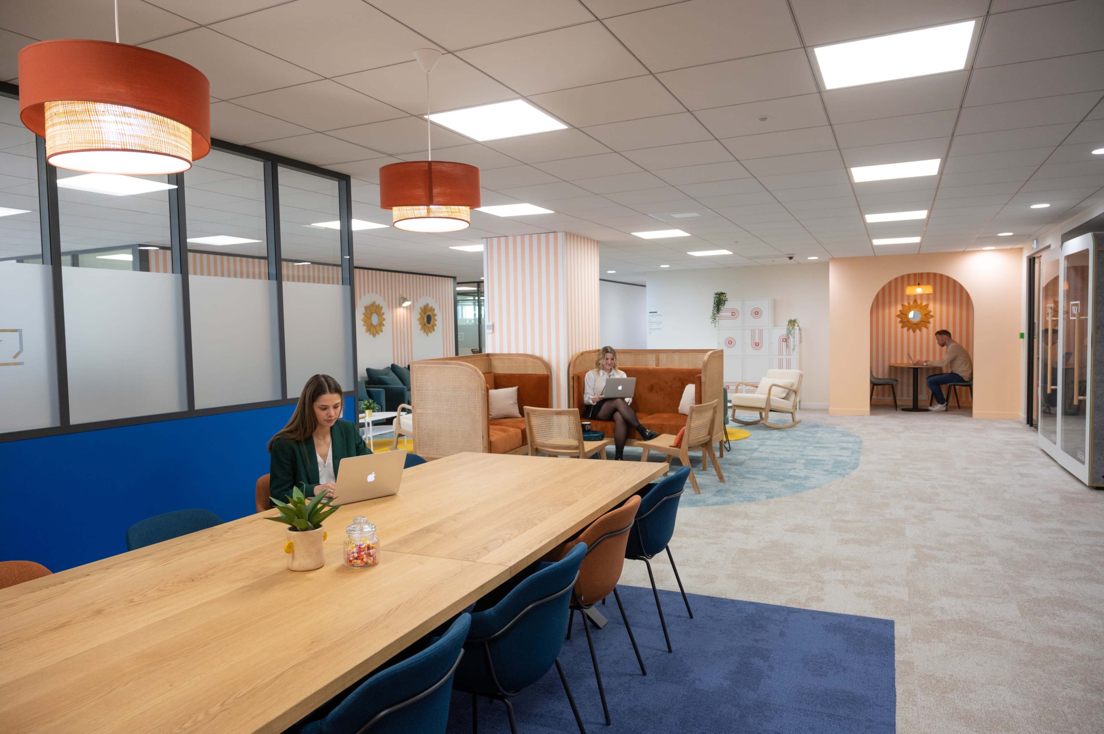 A modern office space featuring a large wooden table, various seating arrangements, and a mix of people working on laptops.
