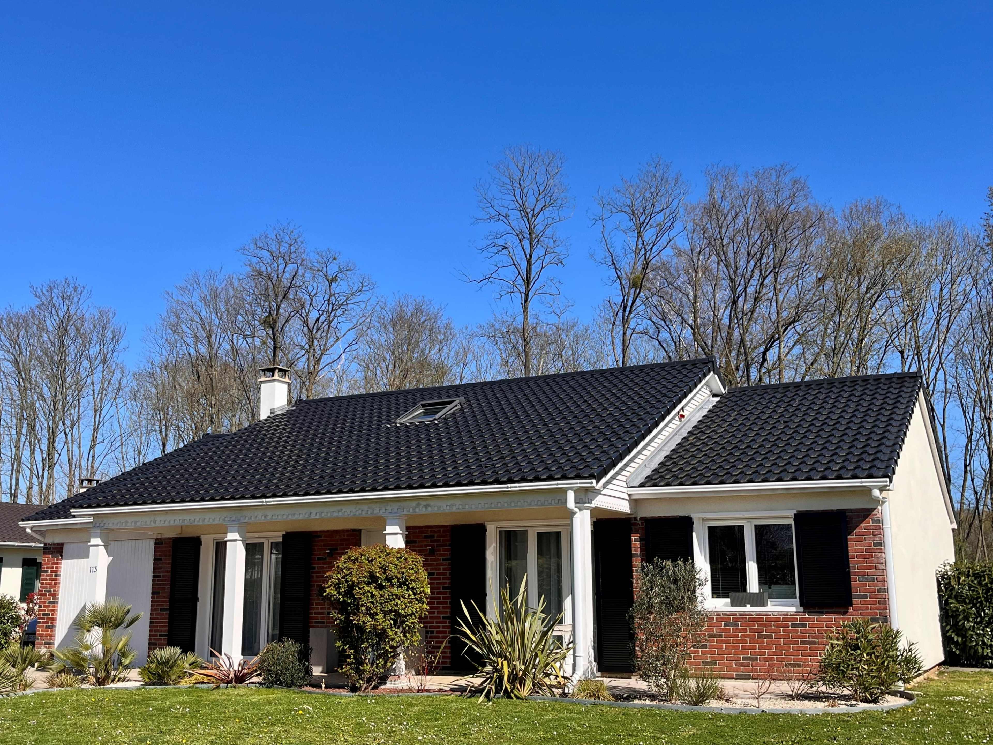 A single-story house with a black roof and brick facade is set against a clear blue sky and surrounded by trees.
