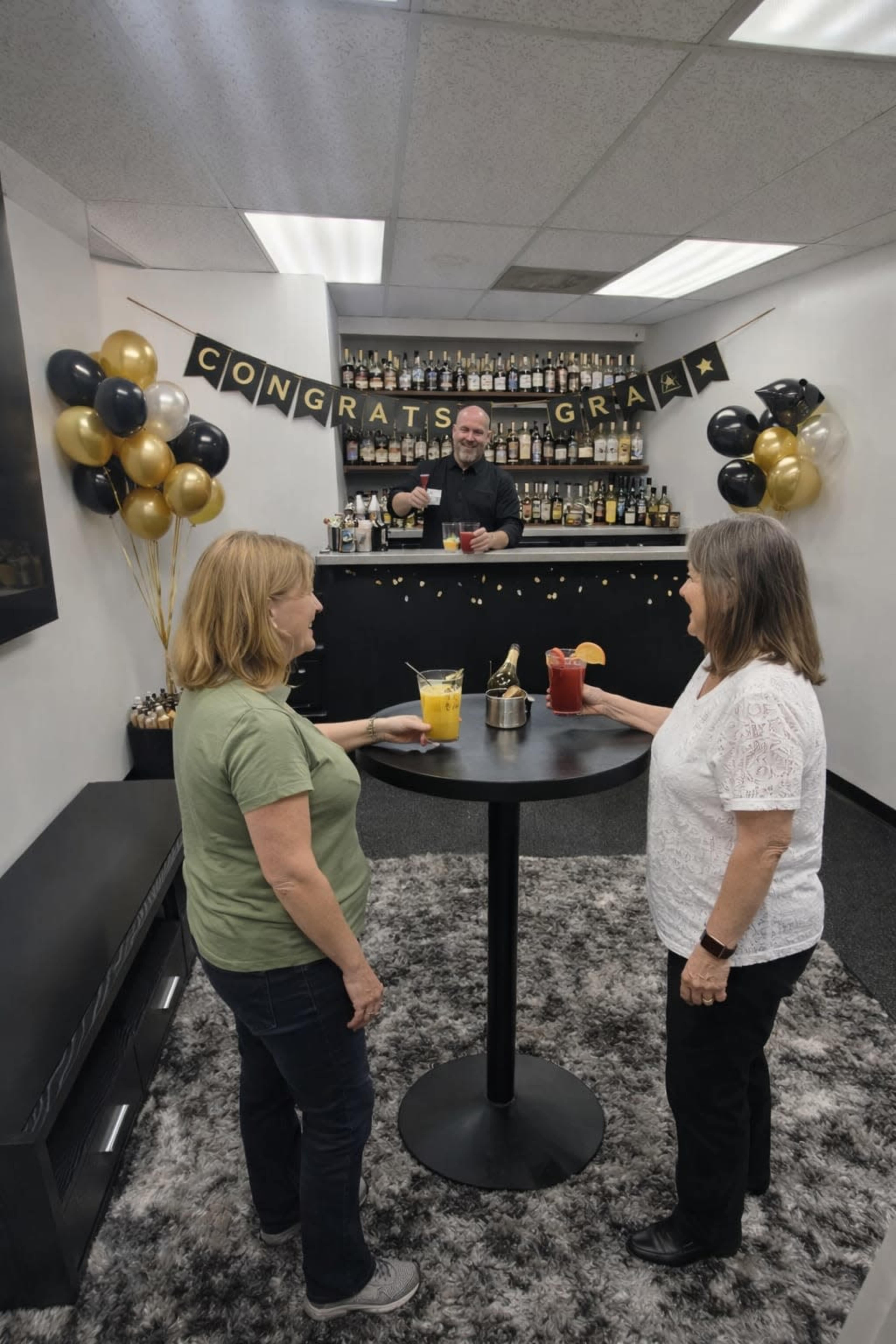 Two women stand at a table in a decorated bar area, while a bartender prepares drinks behind them with "Congrats Grads" banners and festive balloons in the background.