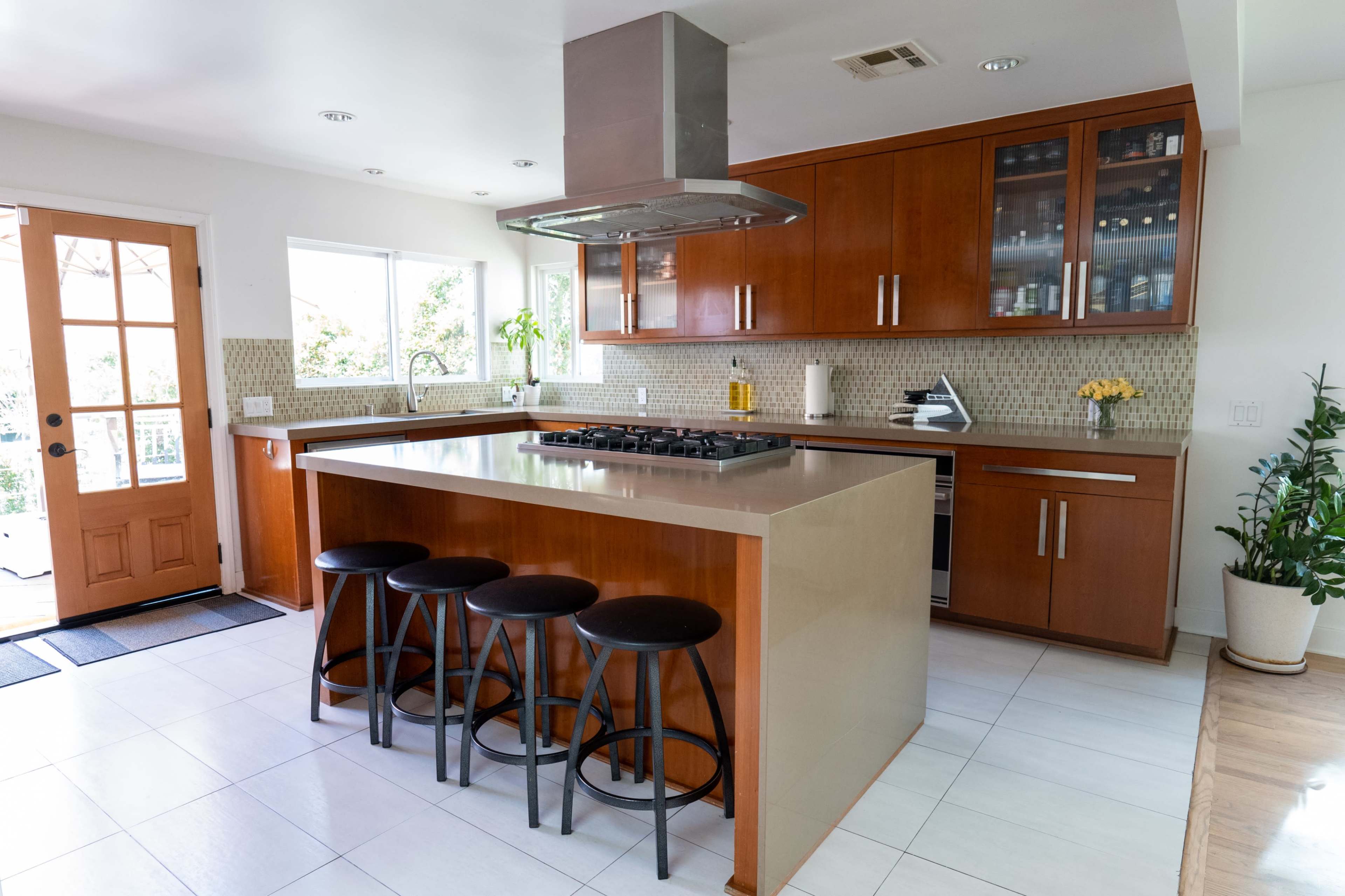 The image shows a modern kitchen with wooden cabinets, a large island with bar stools, and a view of the outdoor area through a glass door.