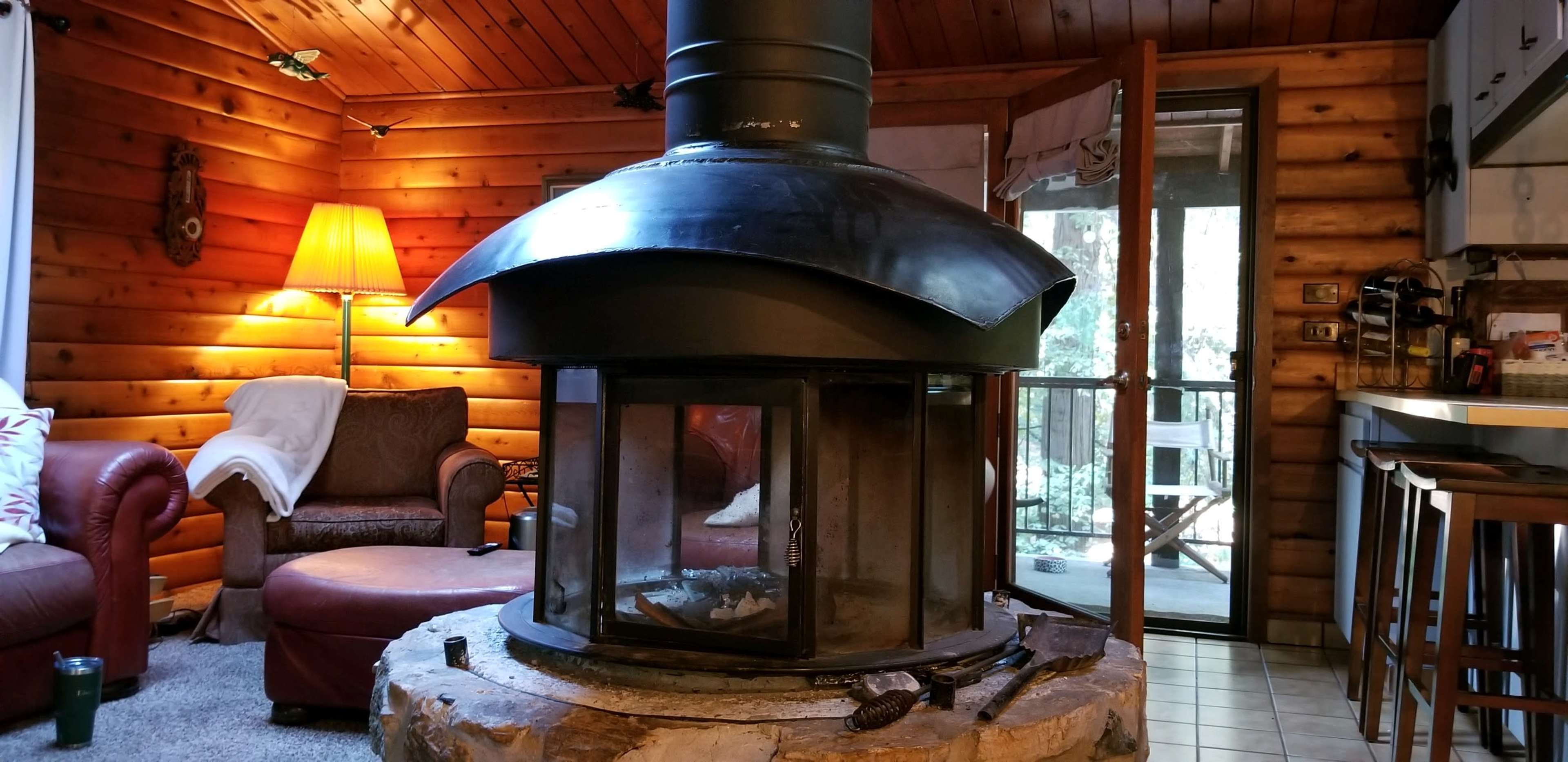 A log cabin interior with a central wood stove, a chair, a floor lamp, and a glass door leading to a balcony.