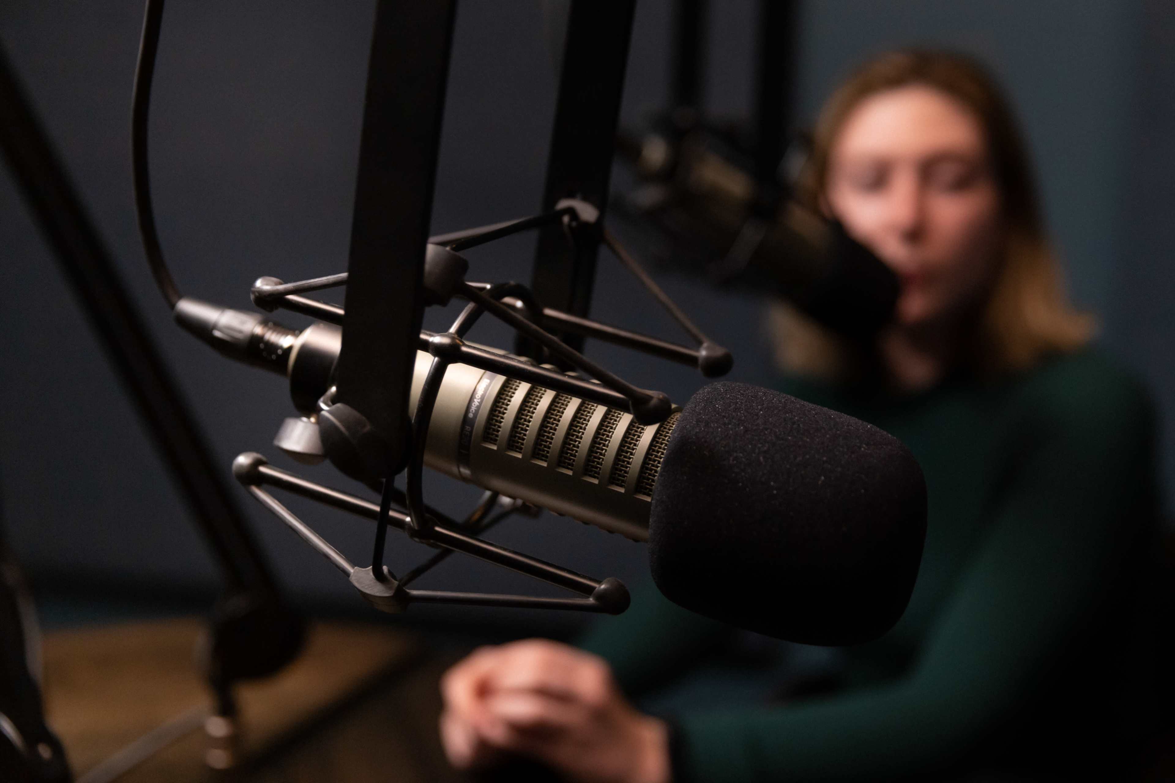 A close-up of a microphone in a recording studio focuses on the device while a blurred figure sits in the background.