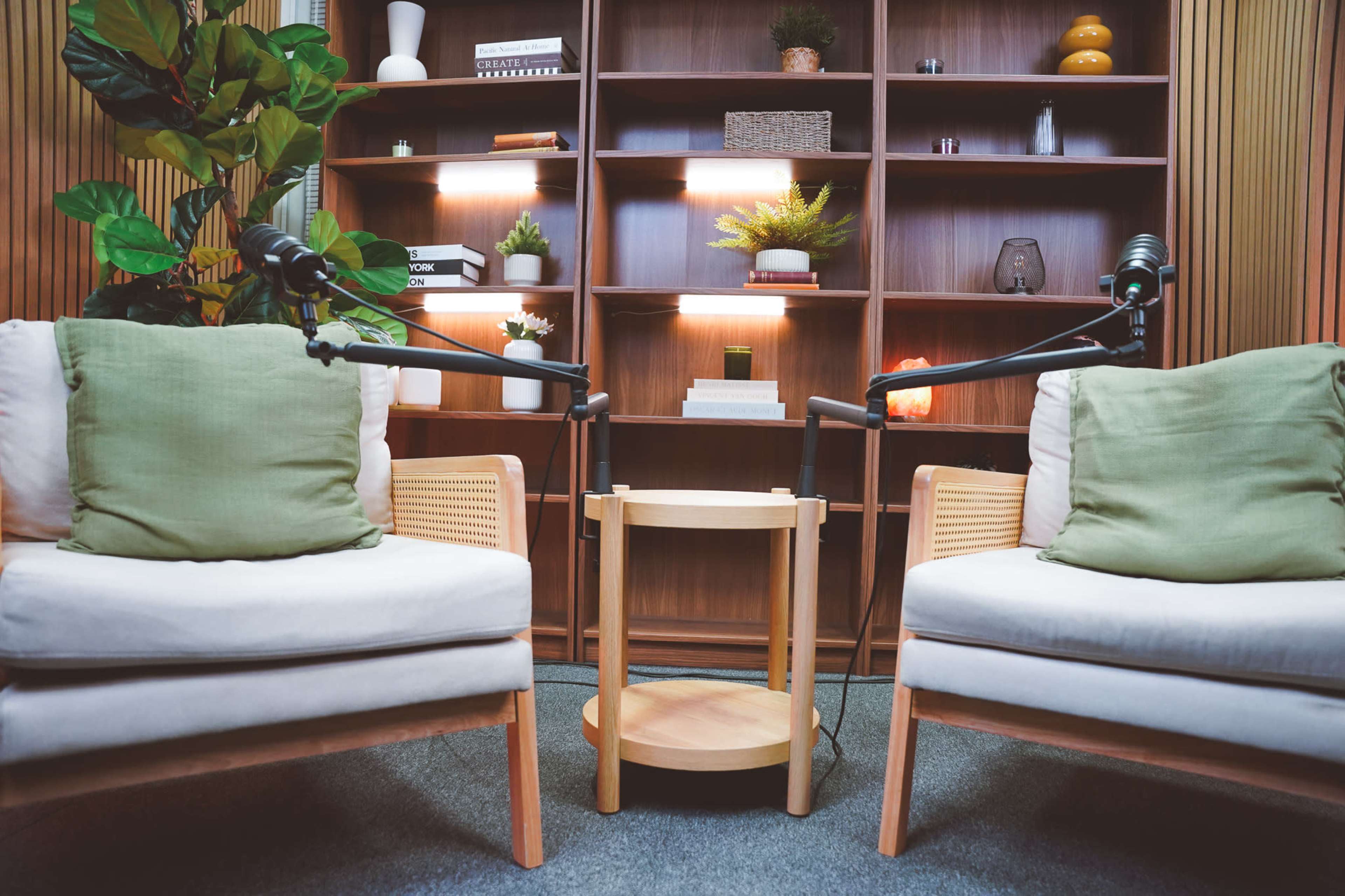 The image shows a cozy seating arrangement with two chairs and a circular table, set in front of a wooden shelving unit filled with plants and decorative items.