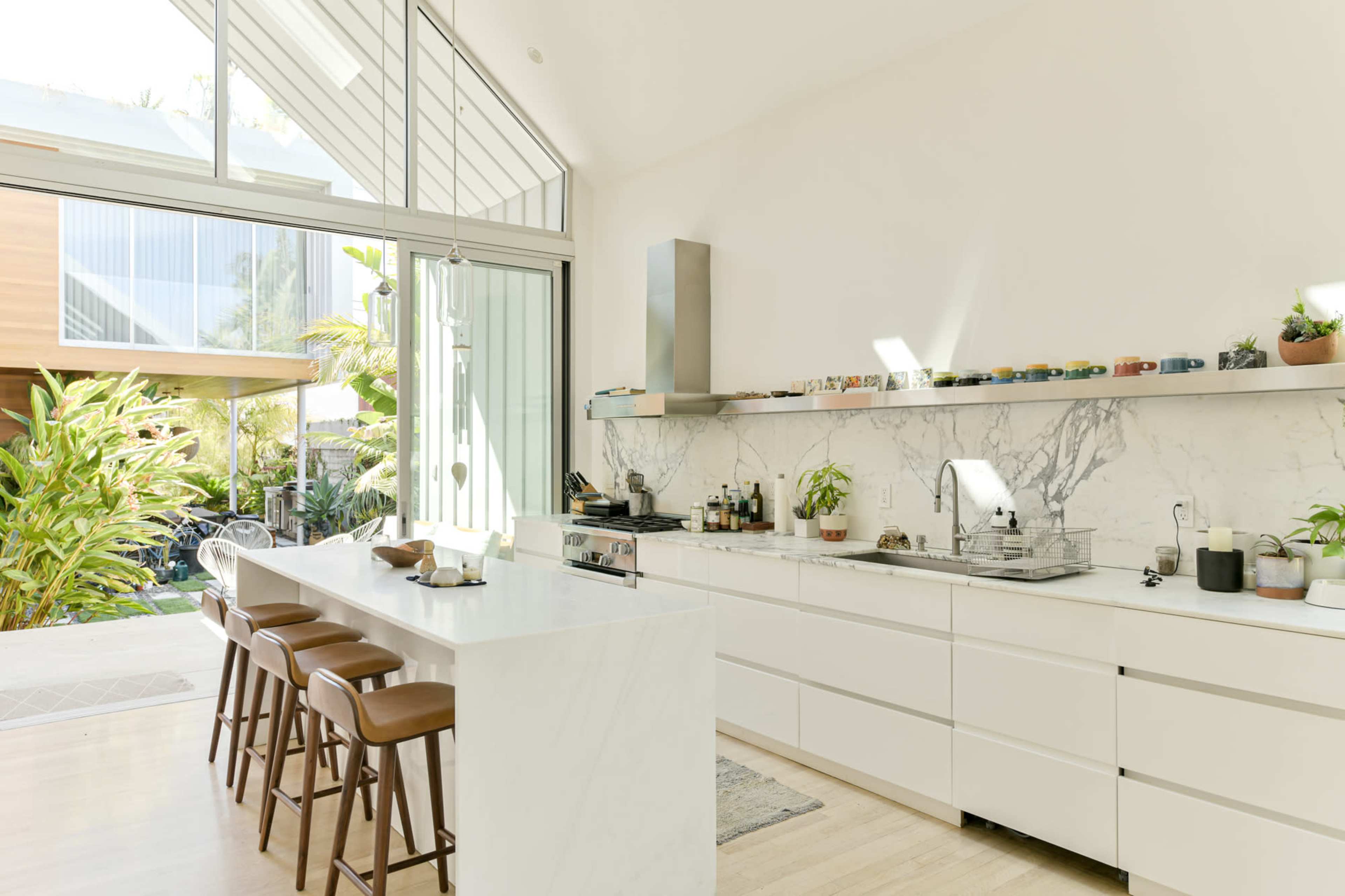 A modern kitchen features a white marble countertop with bar seating and large windows overlooking a lush garden.