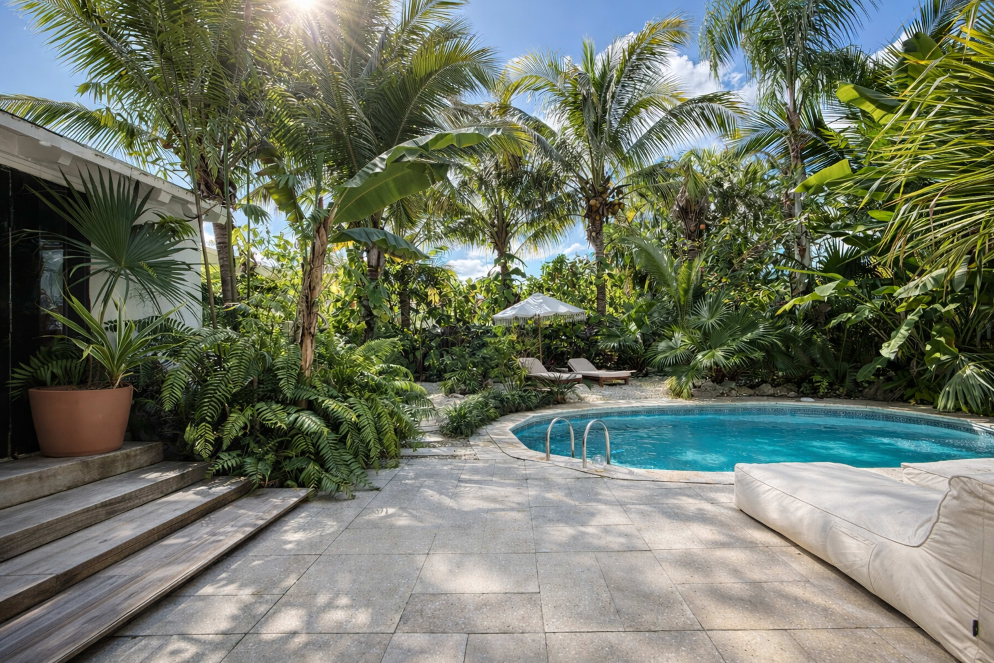 A pool surrounded by lush greenery and palm trees features lounge chairs and an umbrella under a bright sky.