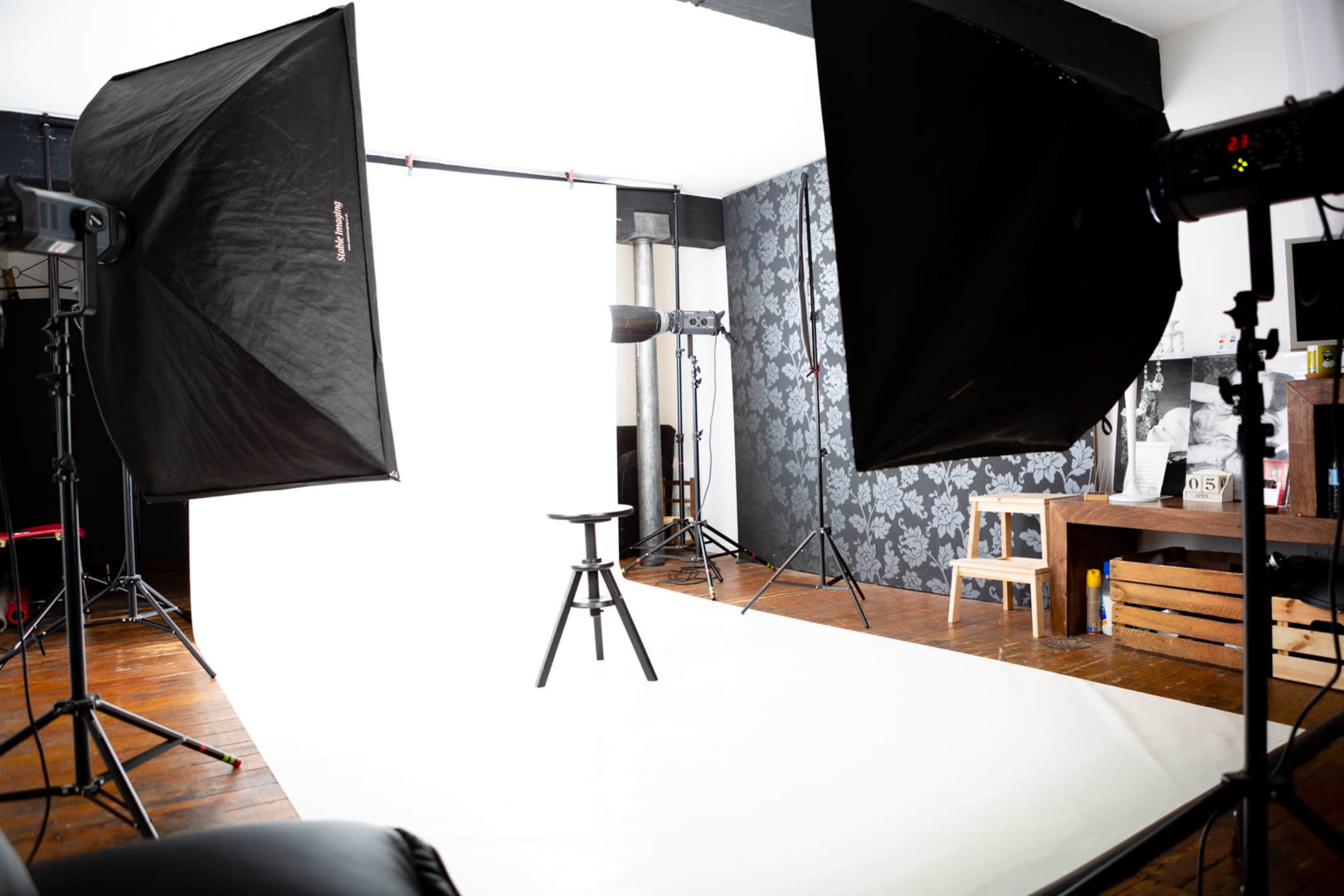 A photography studio setup with a plain white backdrop, a stool in the center, and various lighting equipment positioned around the scene.