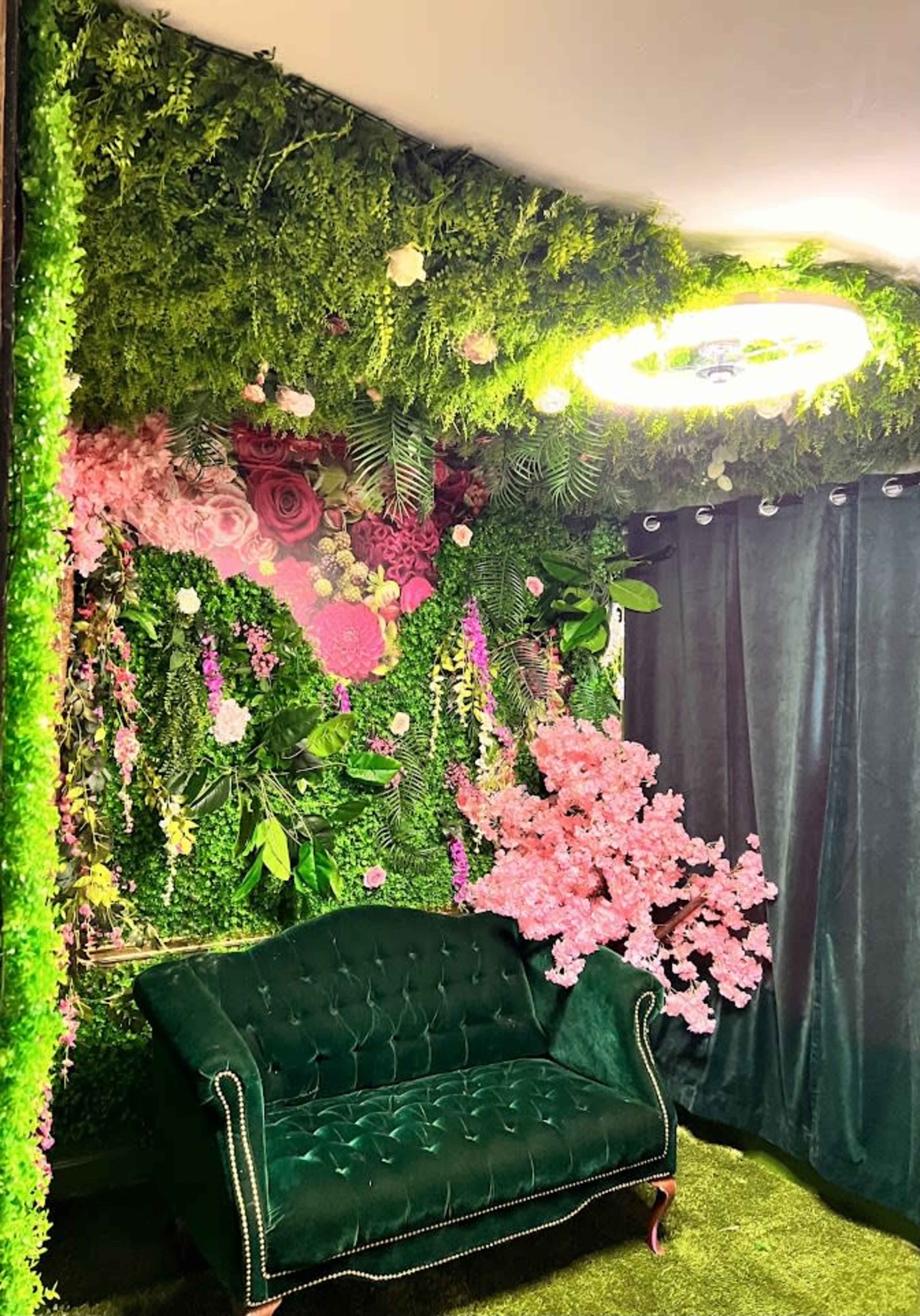 The image shows a room decorated with artificial greenery and flowers, featuring a green velvet sofa in front of a vibrant floral wall.