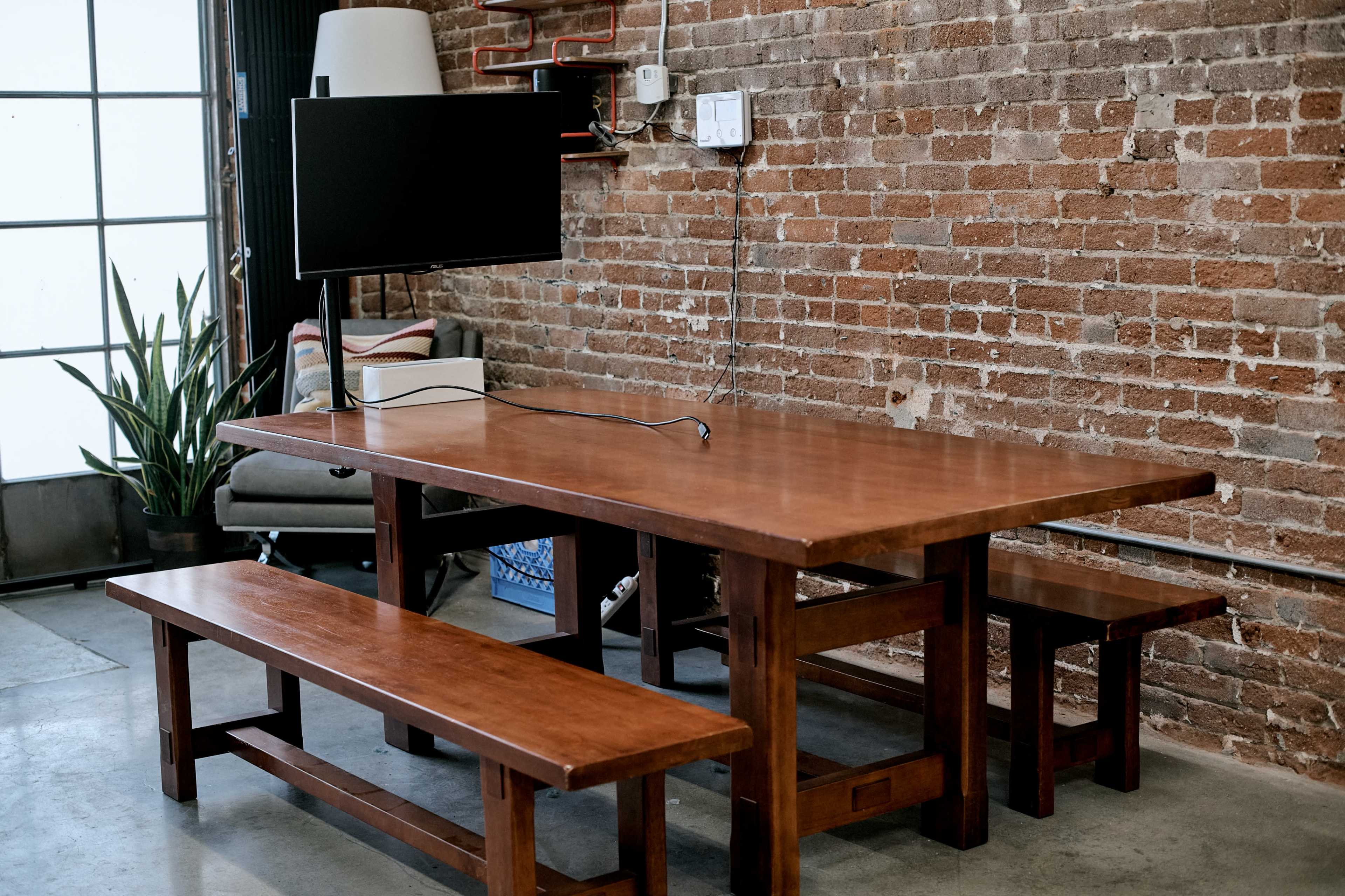 The image shows a wooden table with two matching benches, positioned in front of a television mounted on a wall with exposed brick.
