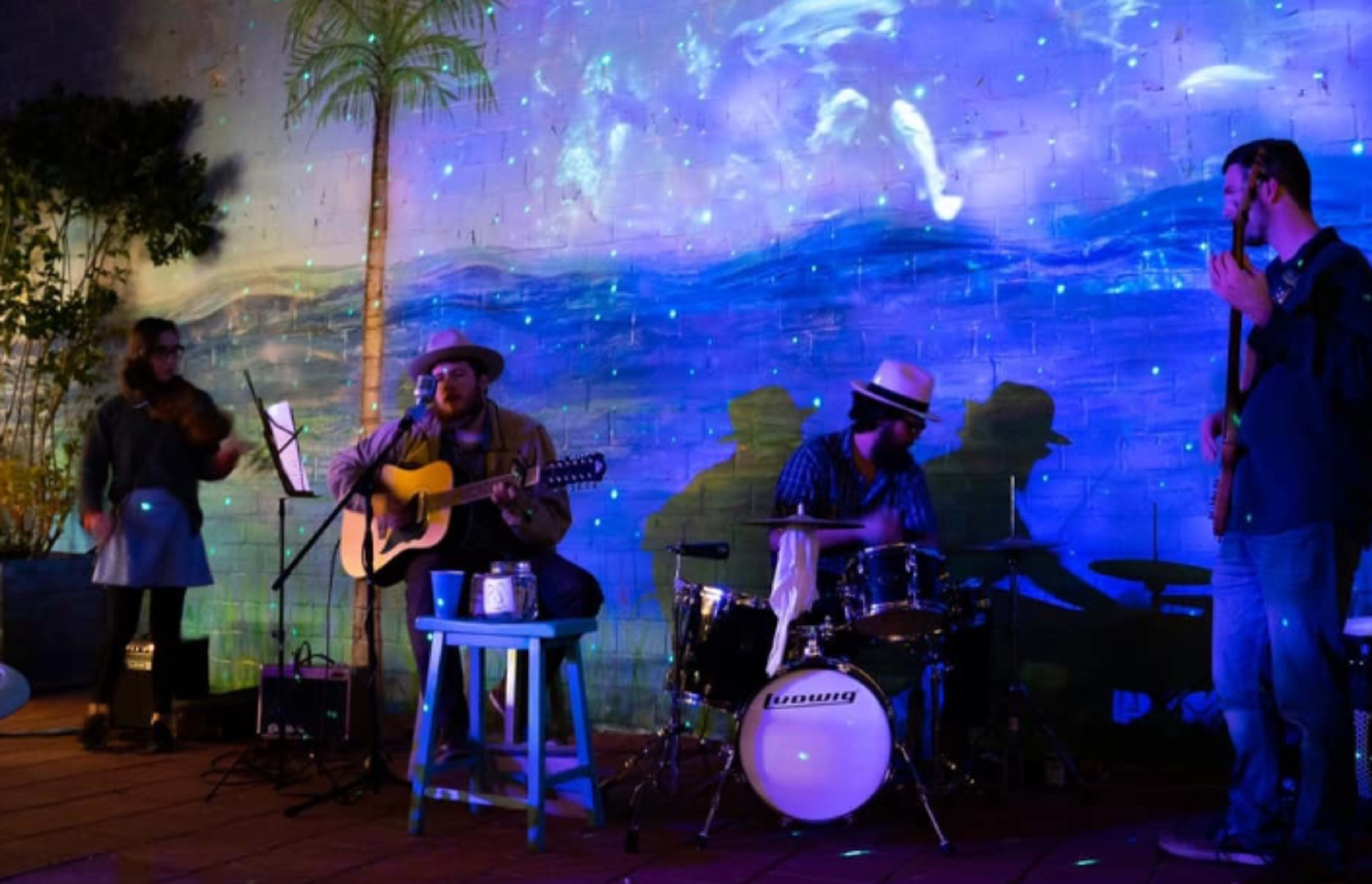 A group of musicians performs on stage in front of a colorful mural while a light display projects shadows of their figures.