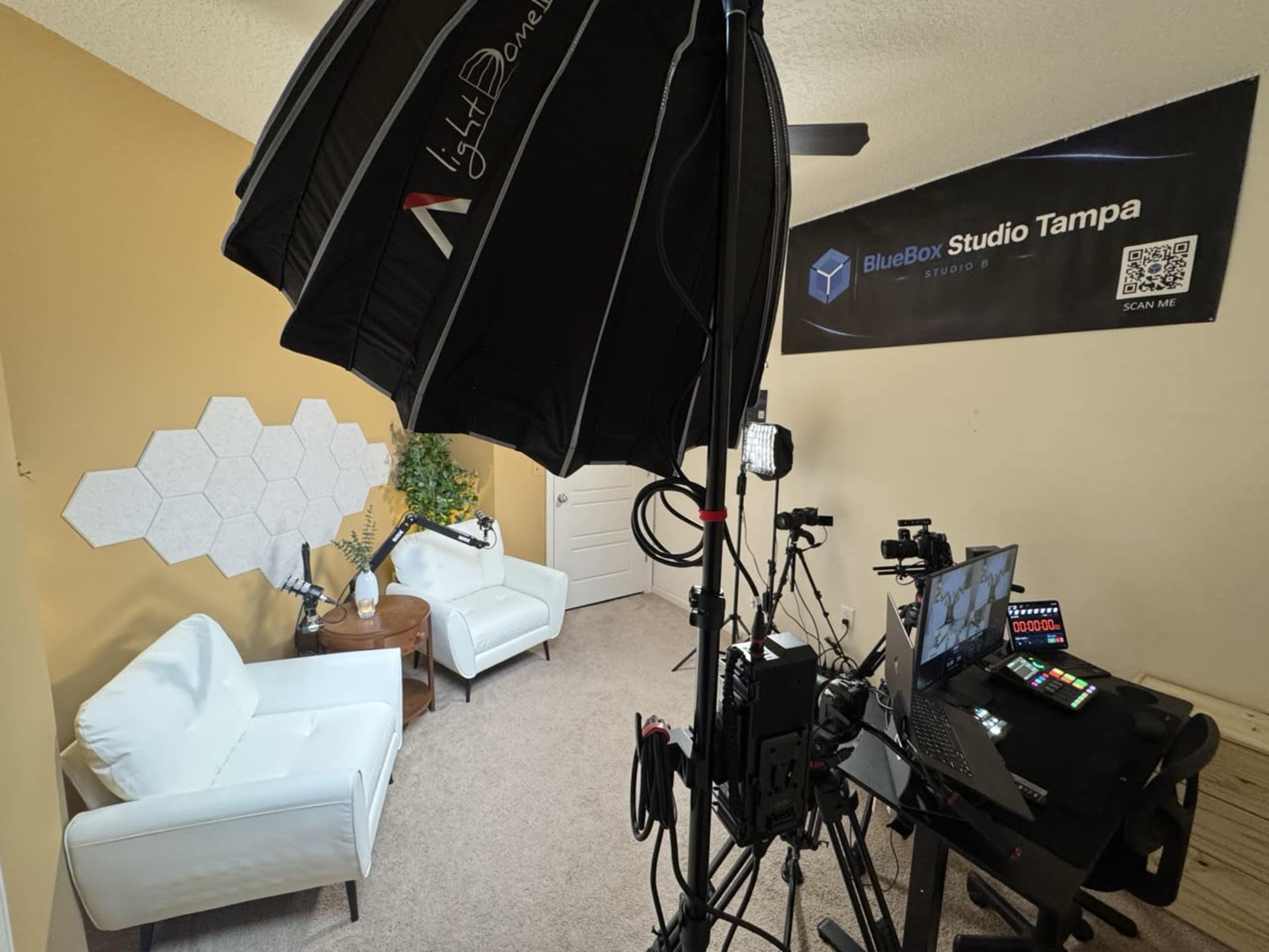 The image shows a recording studio setup featuring two white chairs, a wooden table, a film light, and a computer with various equipment against a yellow wall.
