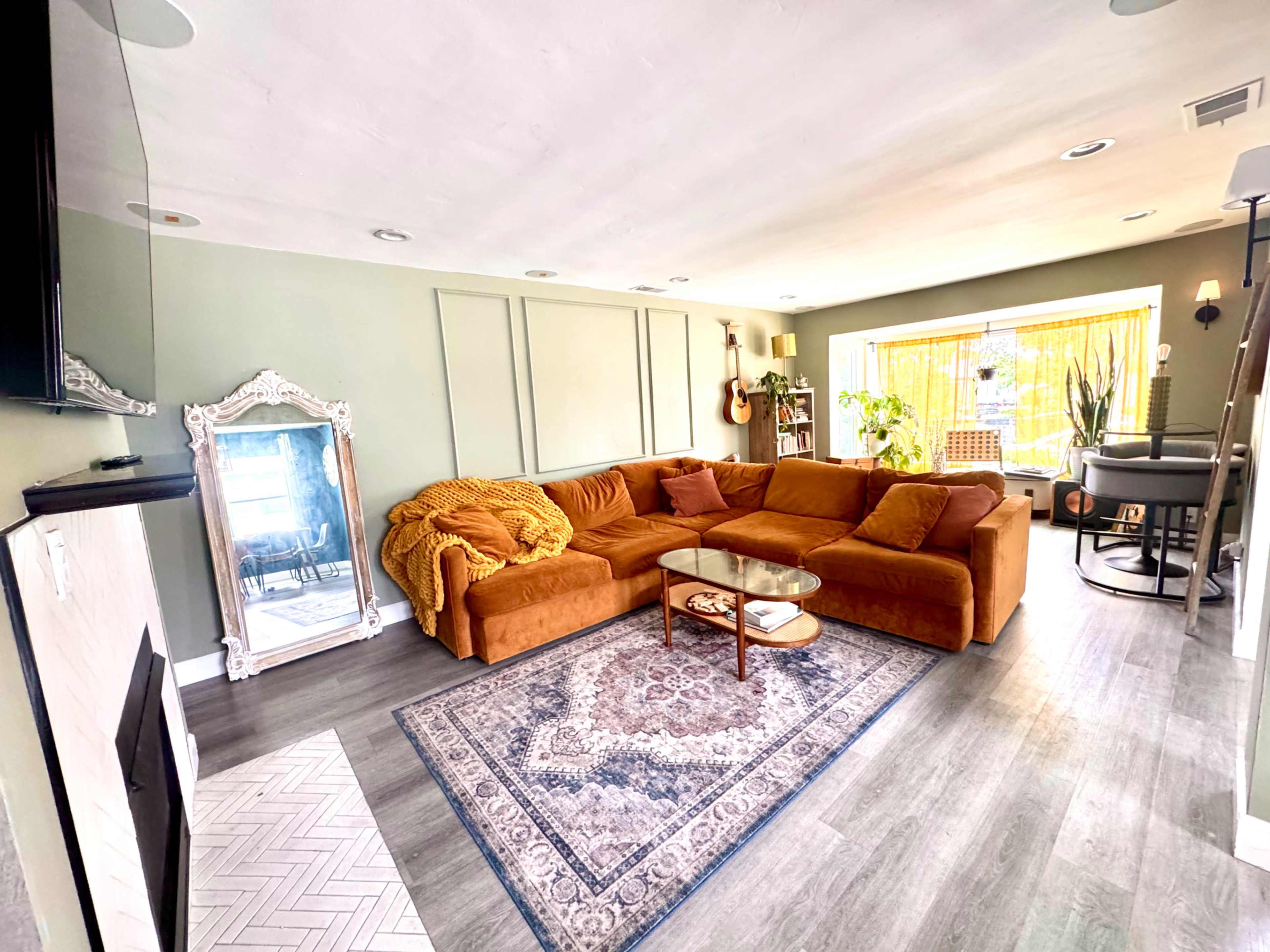 The living room features a large orange sectional sofa, a glass coffee table on a patterned area rug, and large windows with natural light illuminating the space.