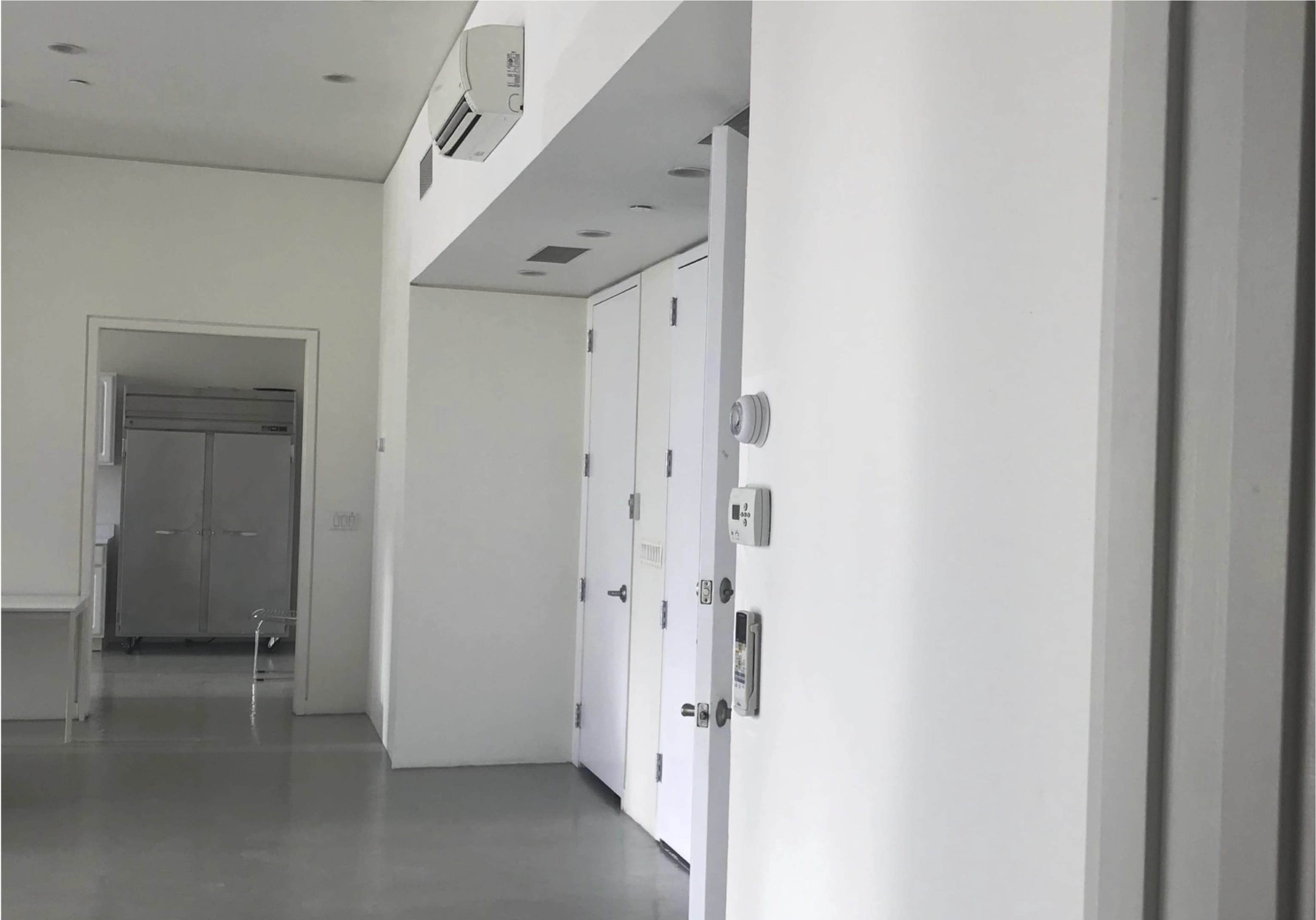 The image shows a minimalist interior with white walls, two closed doors, and a metal kitchen appliance visible in the background.