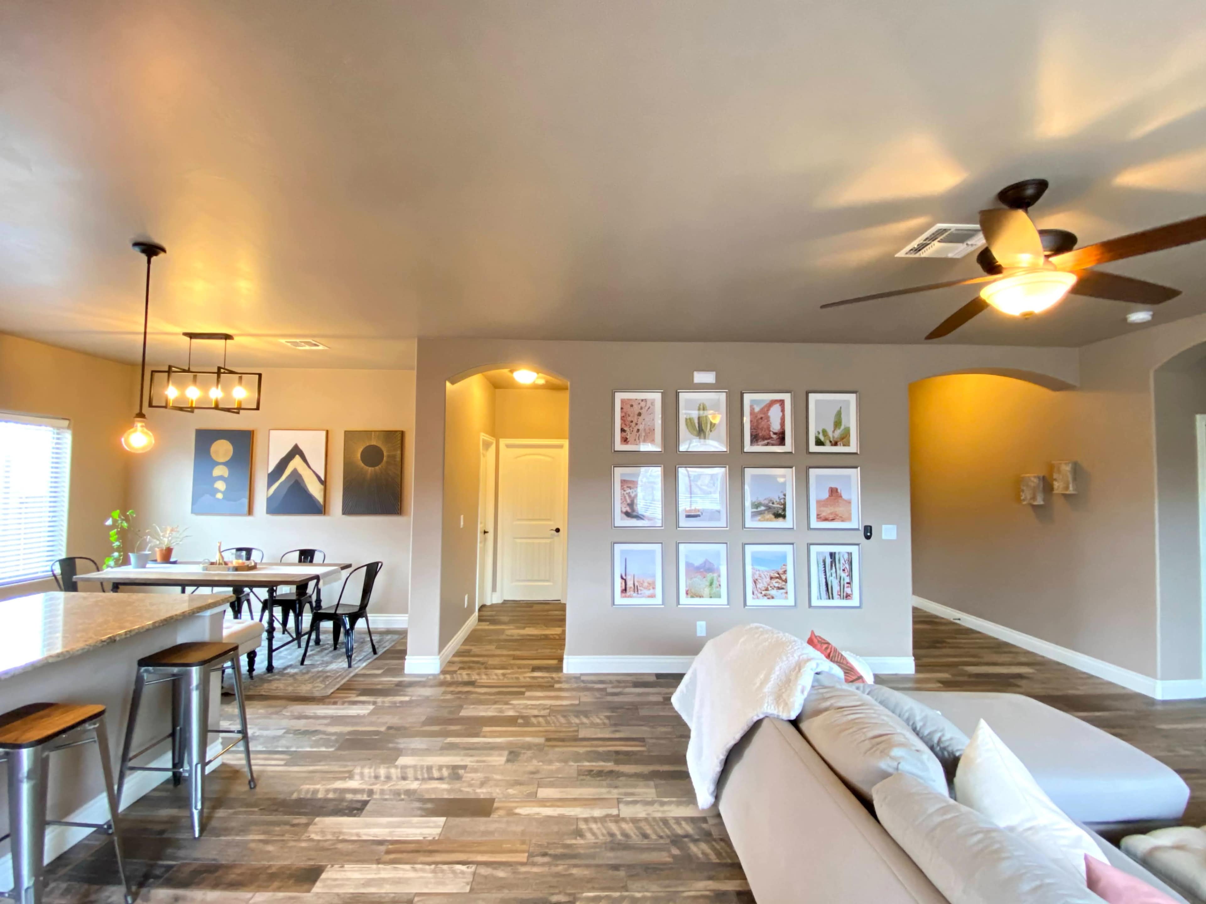 The image shows a spacious living area with a sectional sofa, a dining table, and a gallery wall displaying framed artwork, all set against a neutral wall color and wooden flooring.