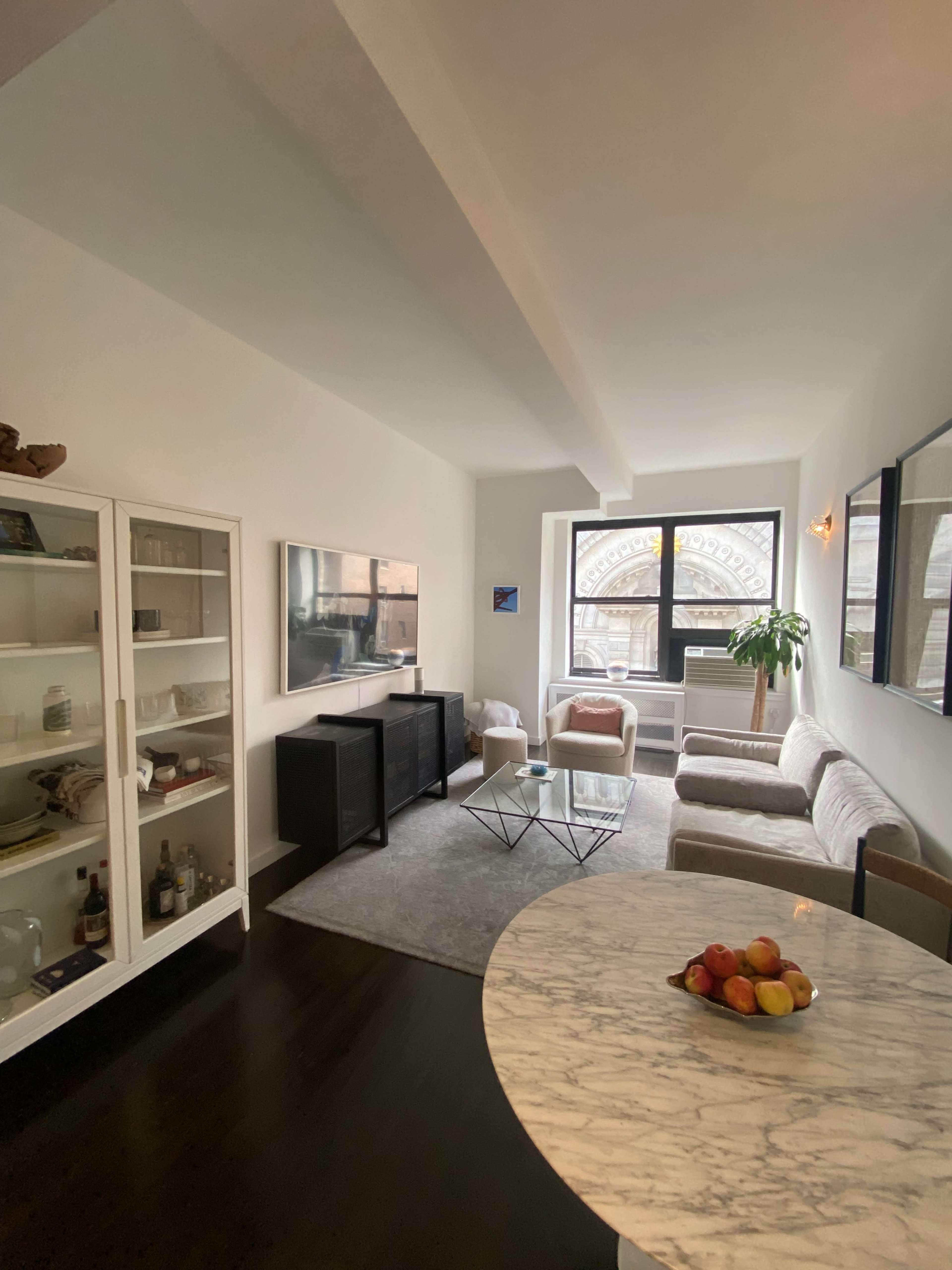 The image shows a modern living room featuring a sofa, coffee table, television, and a nearby dining area with a marble tabletop and a bowl of fruit.