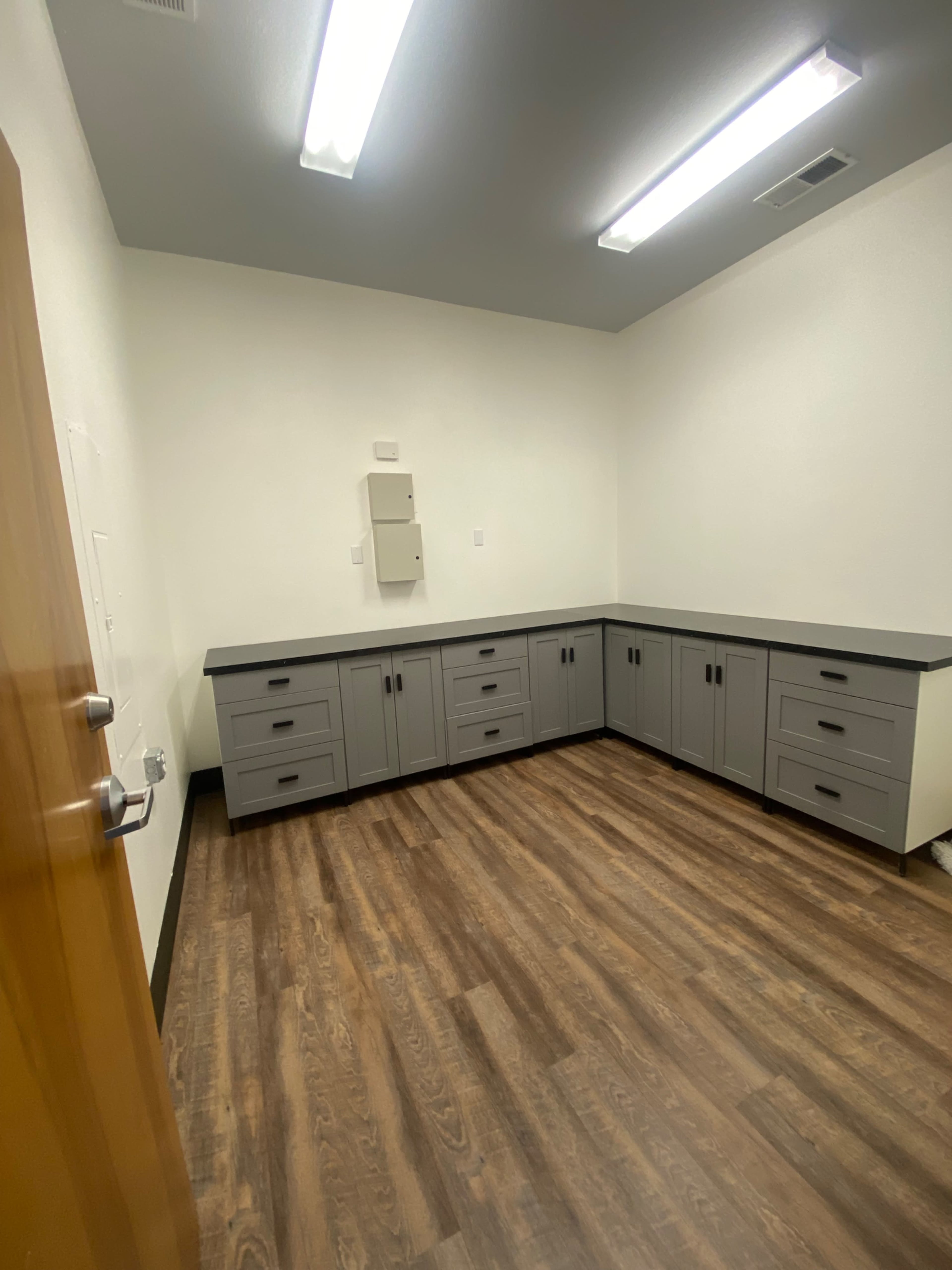 The image shows an empty room with gray cabinetry along two walls and a wood laminate floor, illuminated by overhead fluorescent lights.