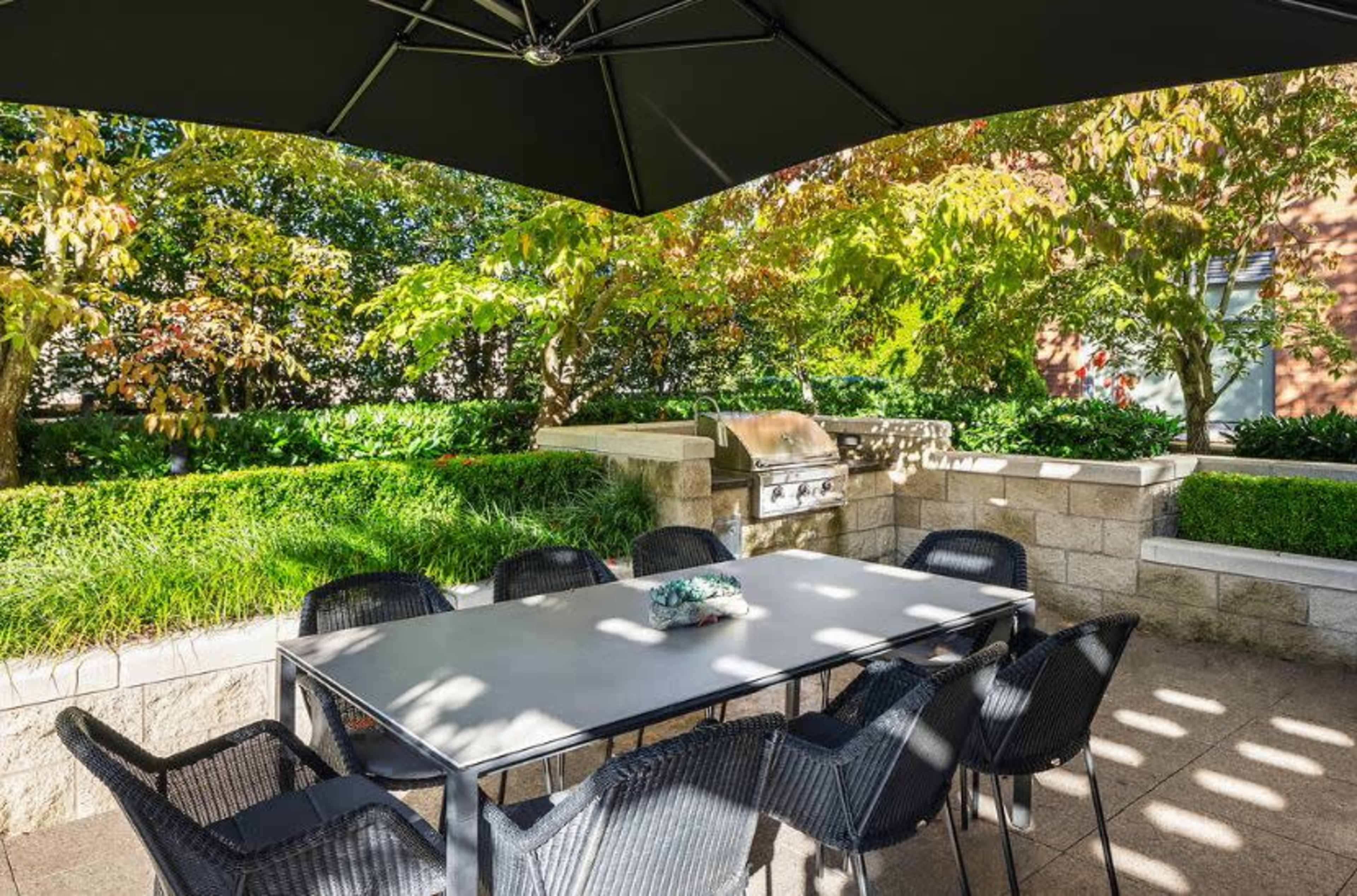A patio area features a large table with chairs under a black umbrella, surrounded by greenery and a stone barbecue grill.