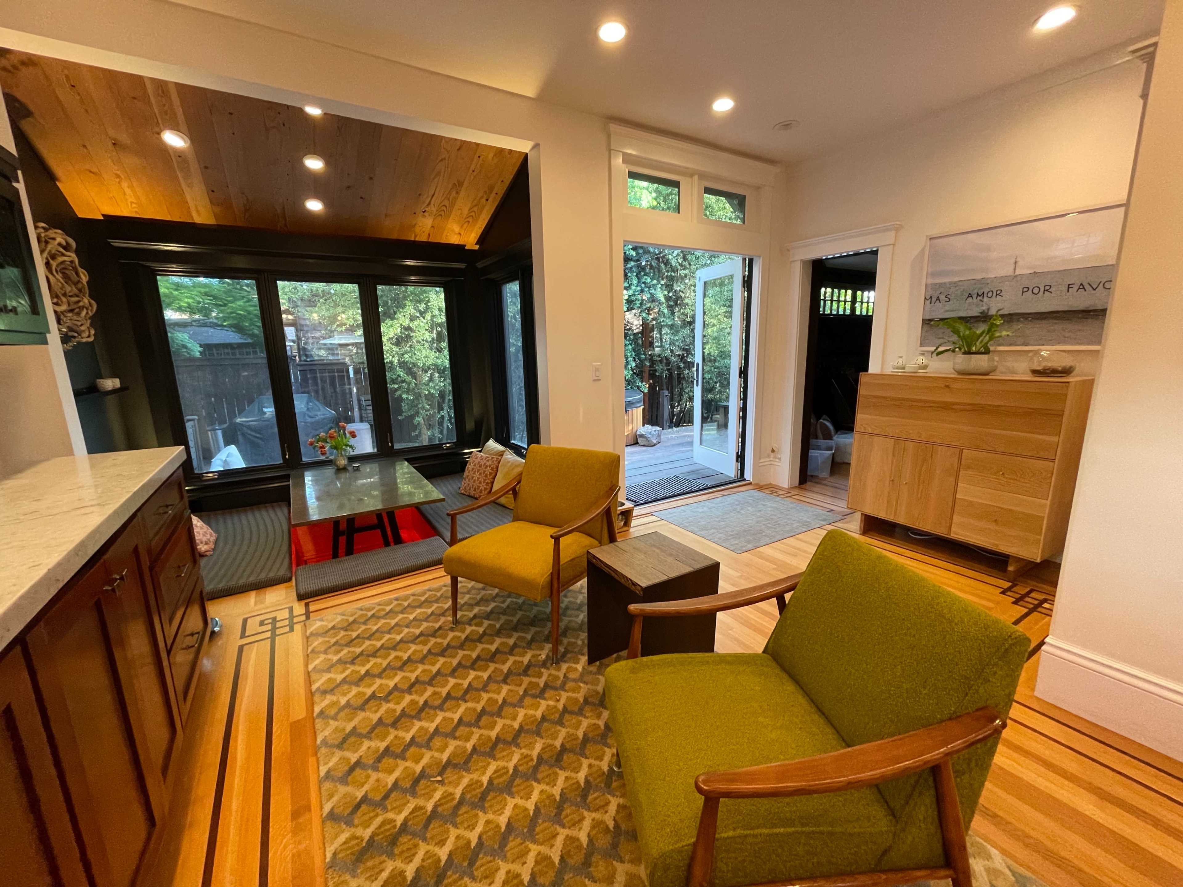 A cozy interior space featuring two green chairs, a wooden table, and large windows that open to a backyard.