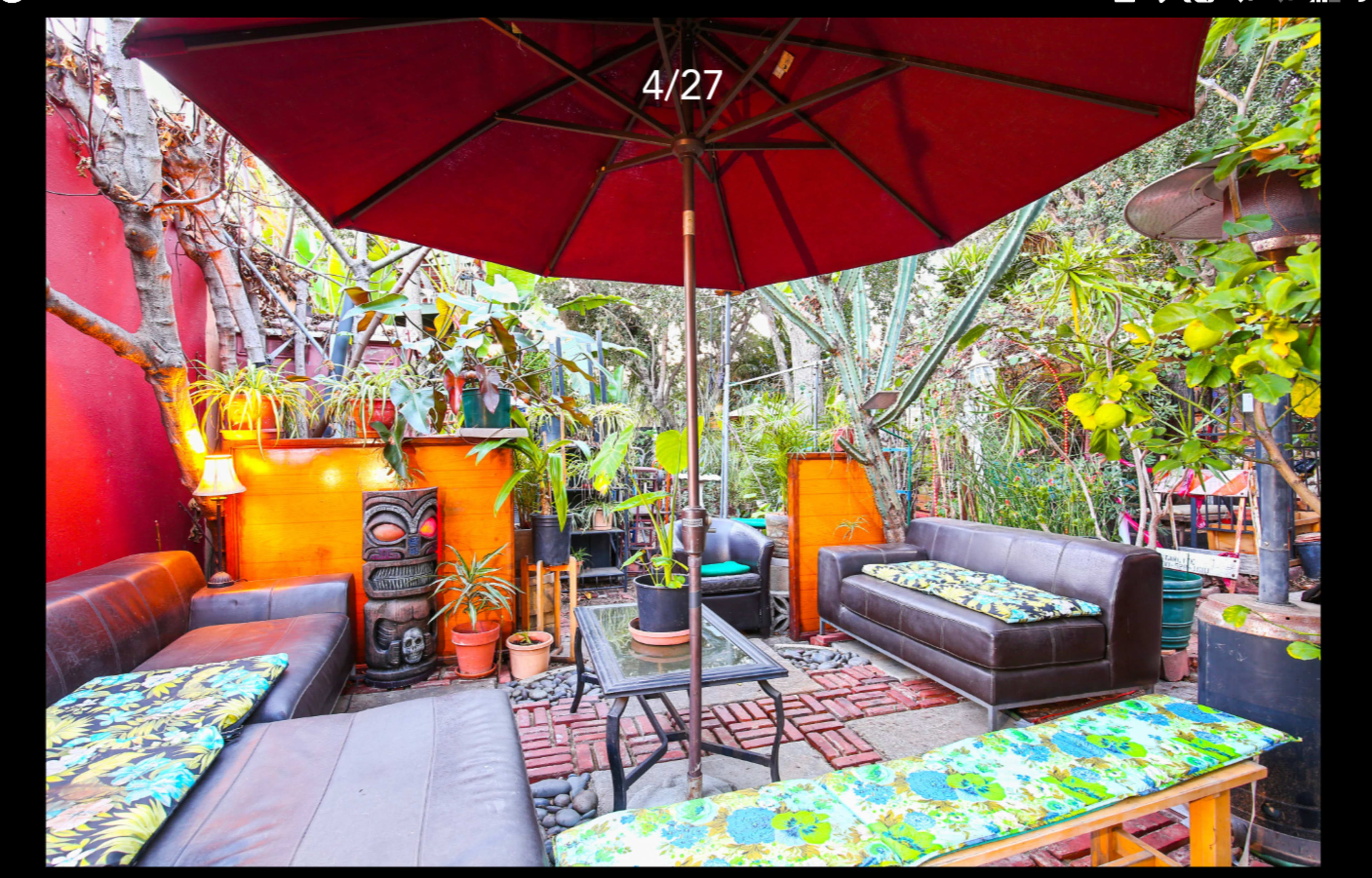 A cozy outdoor seating area features dark brown couches adorned with colorful cushions, shaded by a large red umbrella amidst lush greenery.