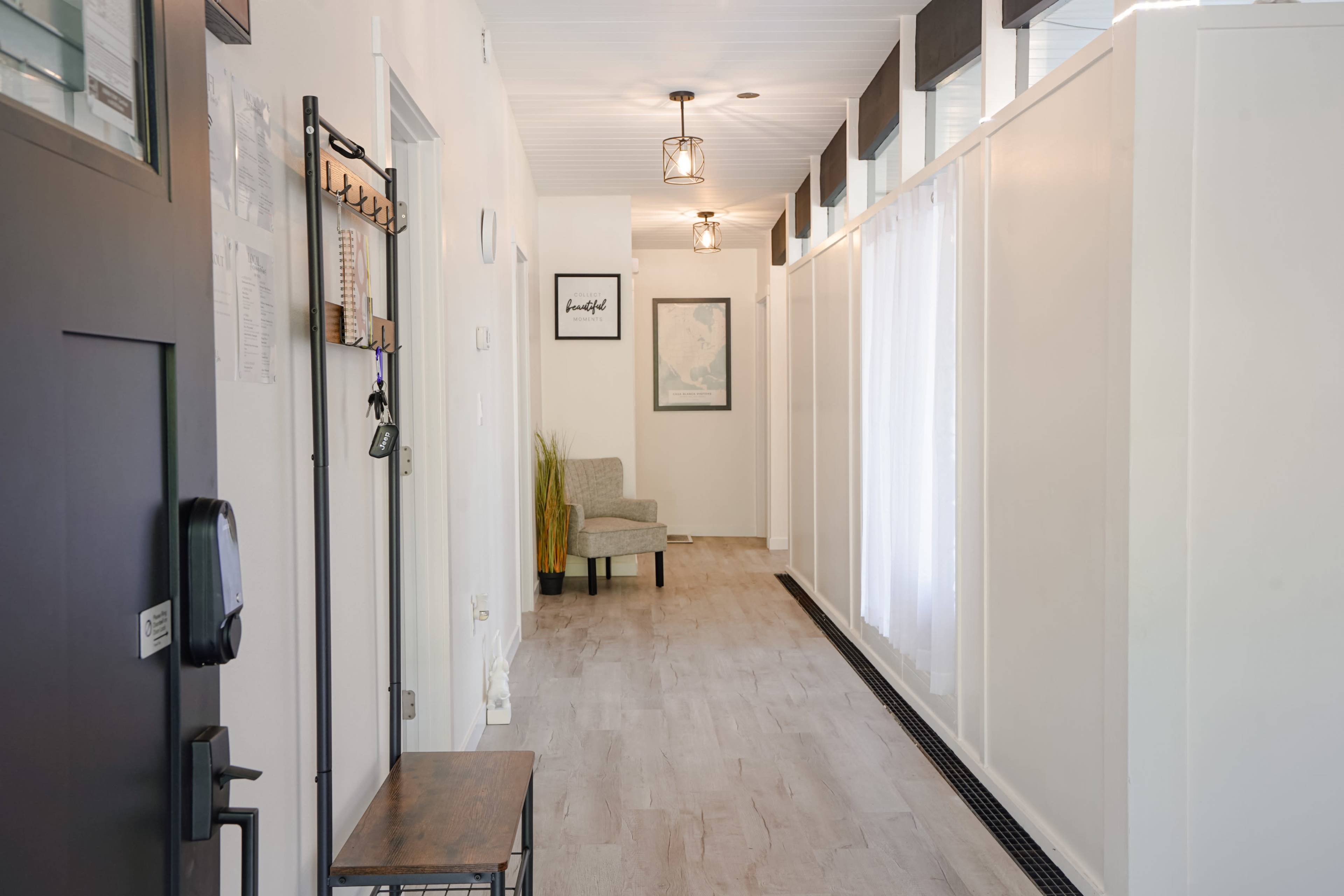 A hallway features a gray chair, decorative plants, wall art, and large windows with sheer curtains.