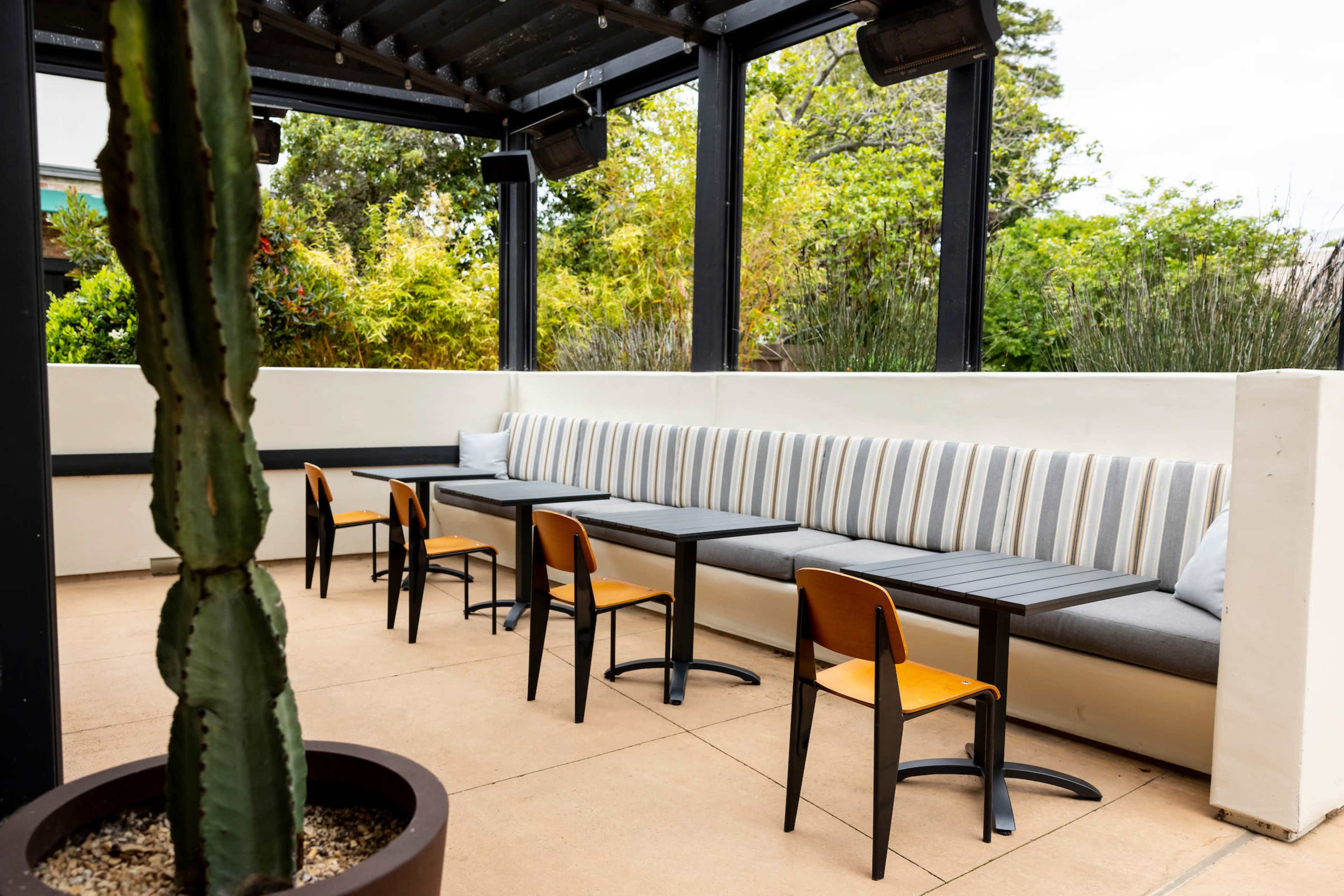 A covered outdoor seating area featuring a long cushioned bench and several black tables with orange-accented chairs, surrounded by greenery.